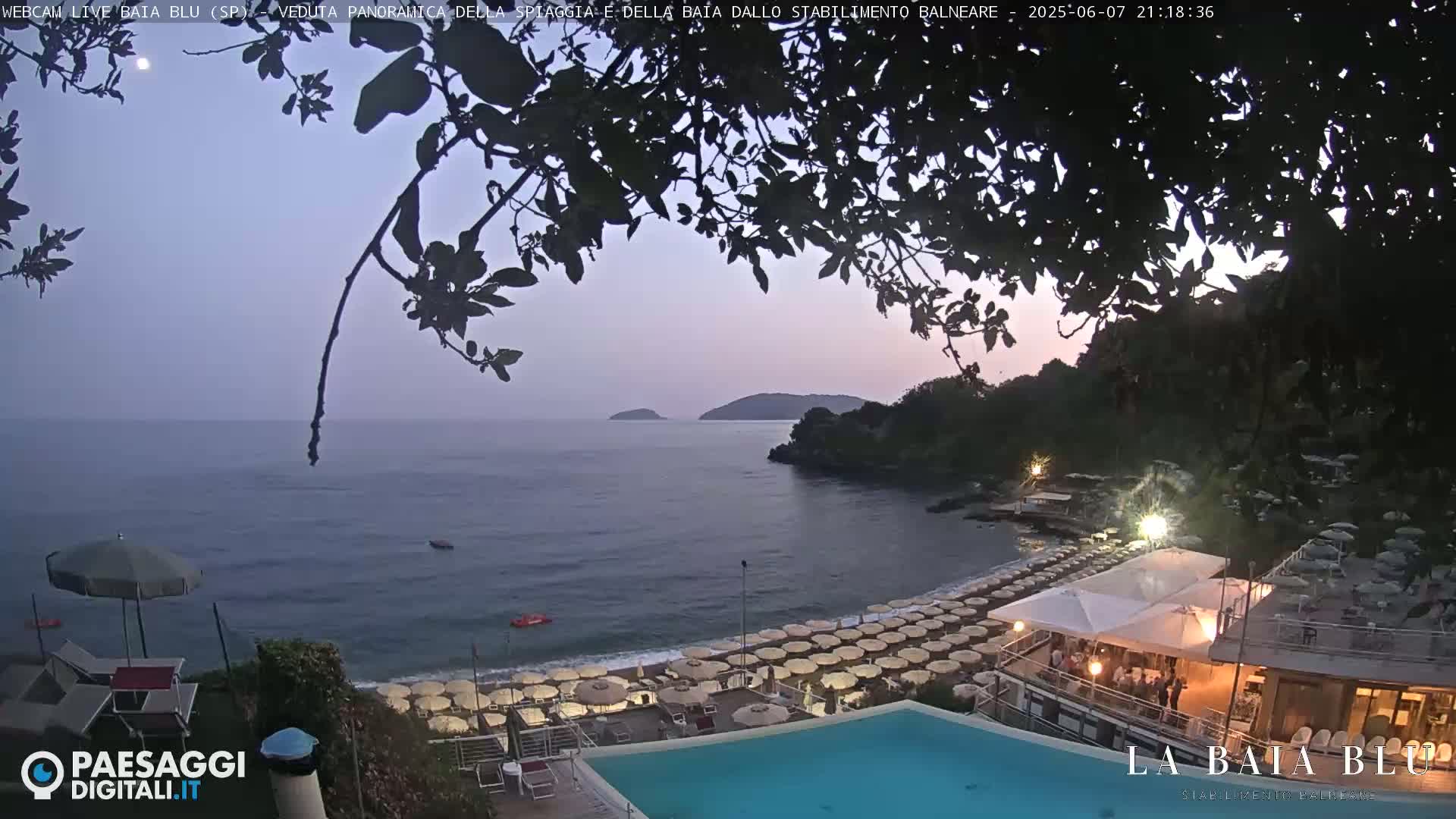 A twilight view of a beach with numerous umbrellas and a swimming pool, overlooking a calm sea with islands in the distance, under a clear sky.