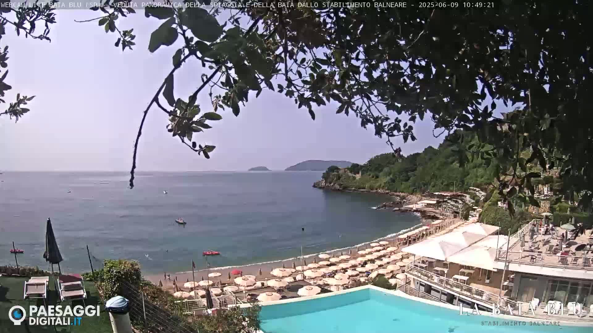 A mostly sunny day overlooking a calm bay with a sandy beach lined with umbrellas, a swimming pool, and buildings on a hillside.