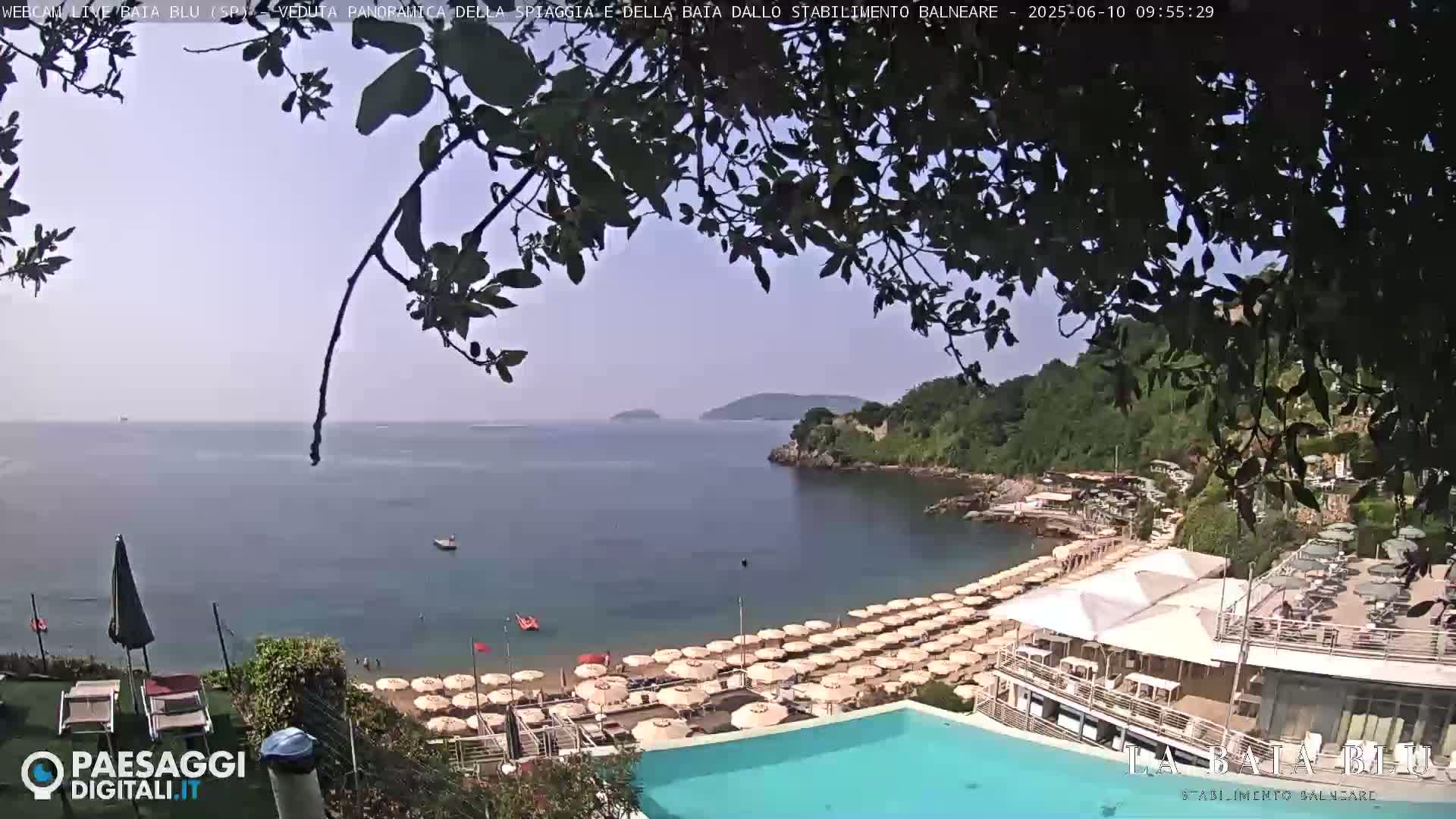A hazy, sunny day overlooking a beach with numerous umbrellas and a swimming pool, situated on a calm bay surrounded by green hills.
