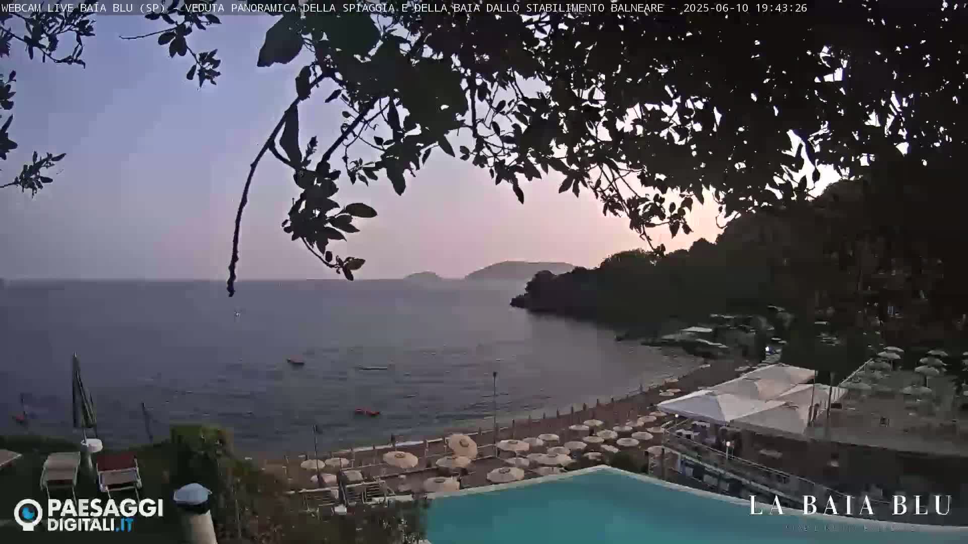 A beach with numerous umbrellas and a swimming pool is situated on a calm, twilight-lit ocean cove, partially obscured by overhanging tree branches.