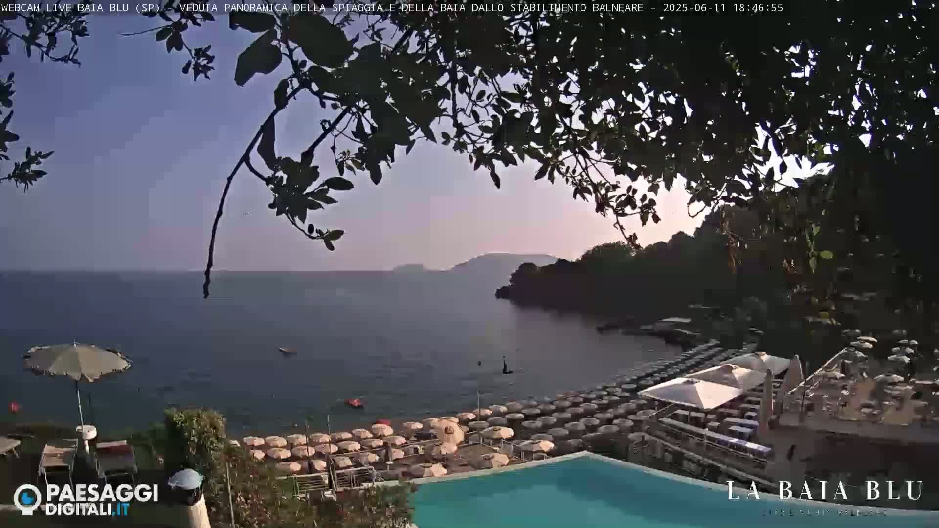 A tranquil beach scene at sunset shows a calm body of water with numerous beach umbrellas and a swimming pool, partially obscured by tree branches.
