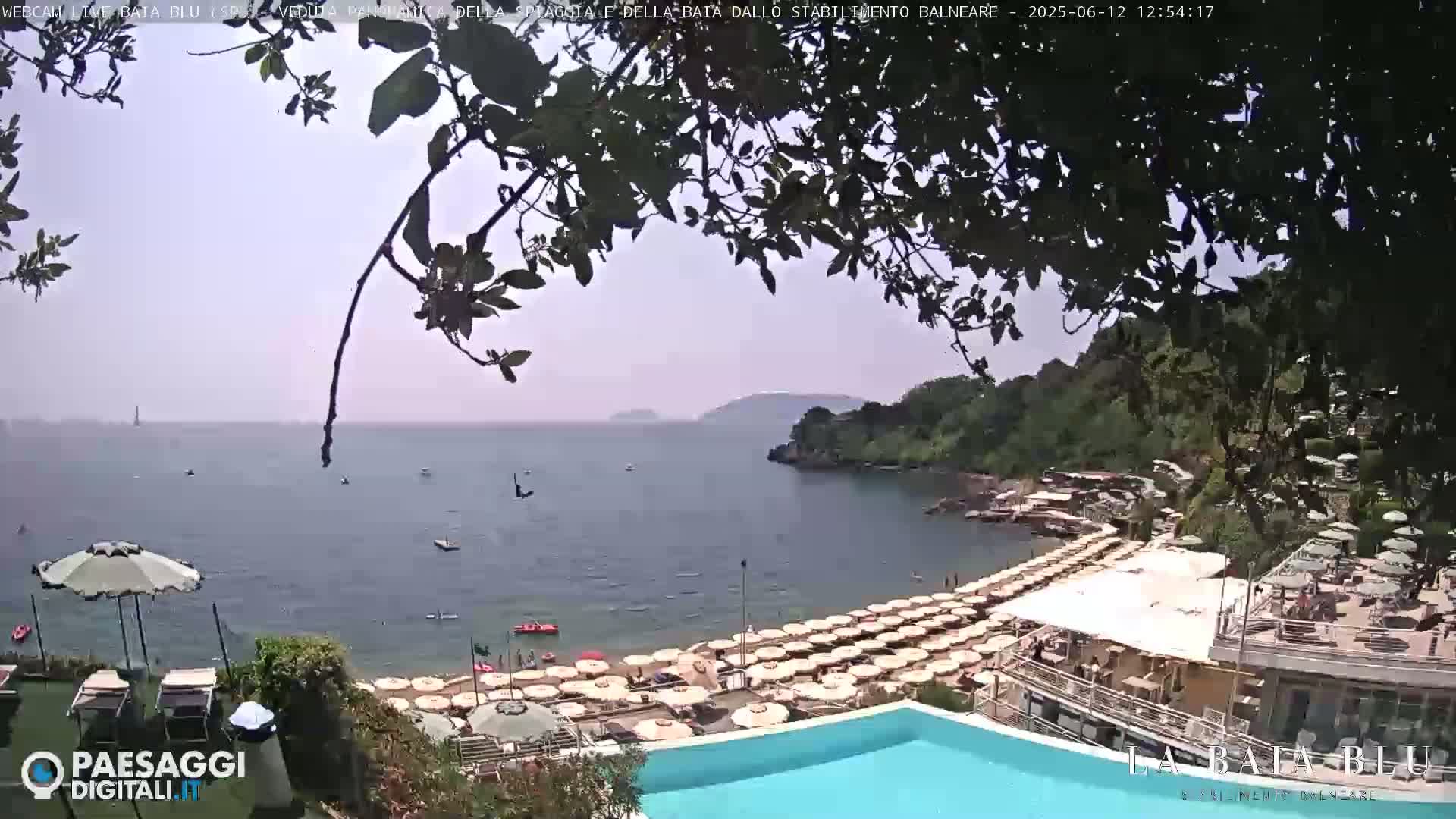 A hazy sunny day overlooks a calm bay with a beach lined with umbrellas and lounge chairs, a swimming pool, and some buildings.