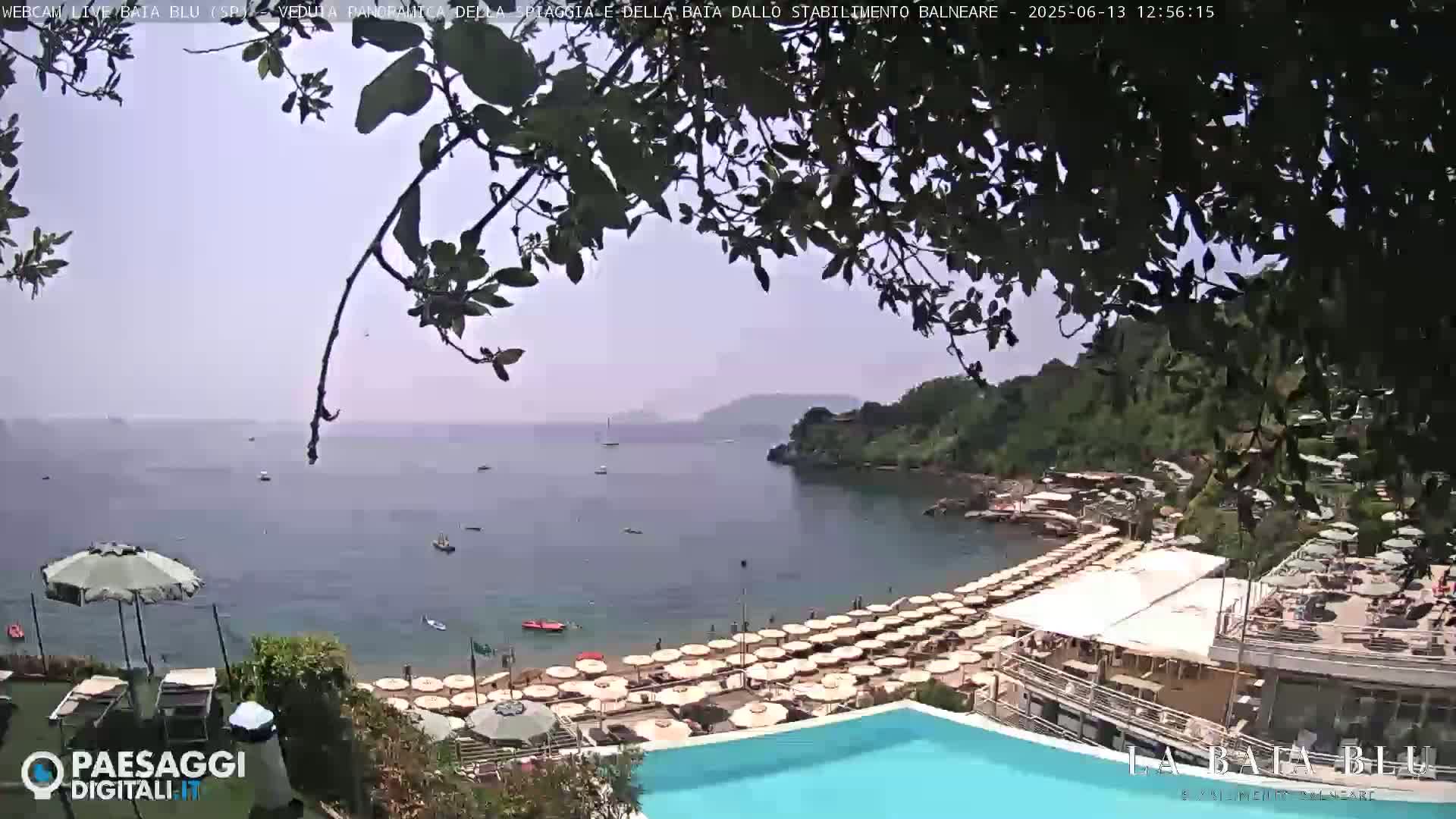 A hazy, sunny day overlooks a calm bay with a beach full of umbrellas, a swimming pool, and a few boats in the distance.