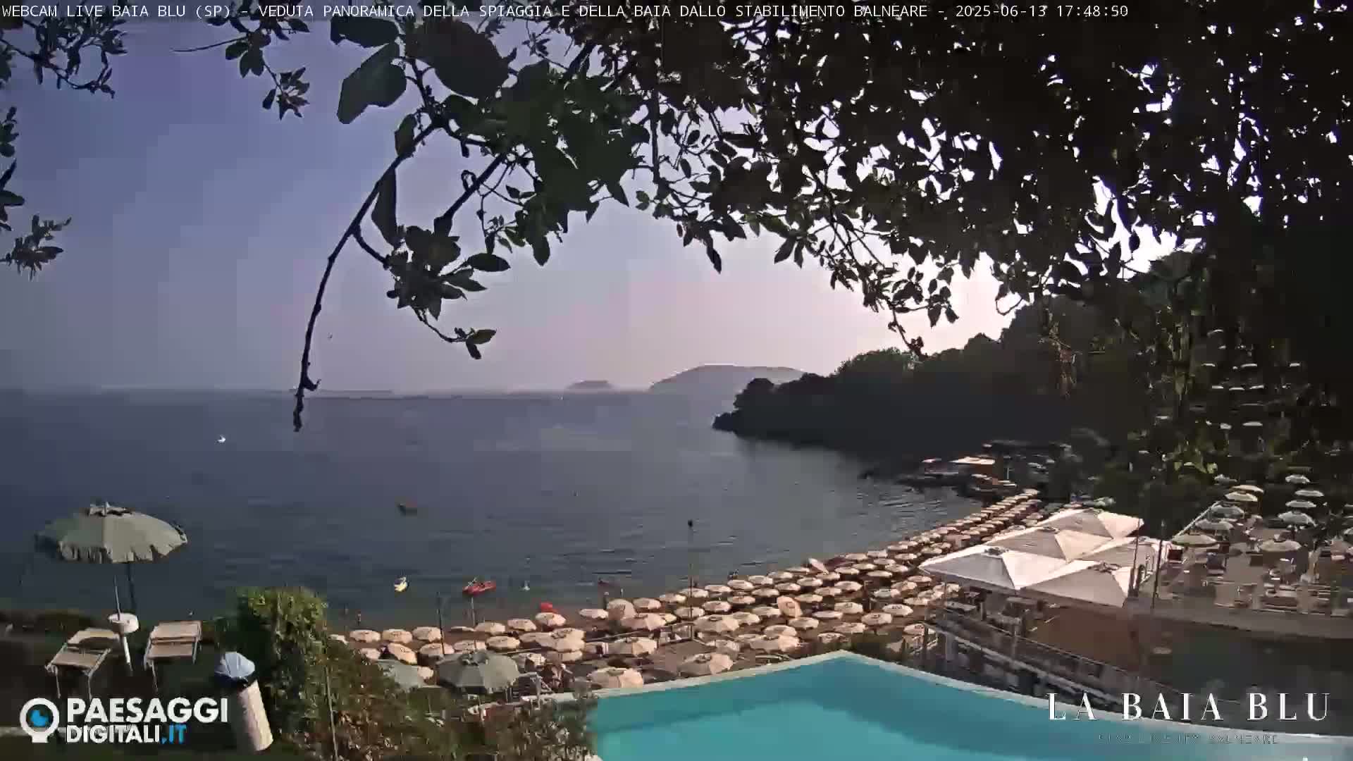 A partly sunny view from a high vantage point shows a calm bay with a beach full of umbrellas, a swimming pool, and some green trees, partially obscuring the view.