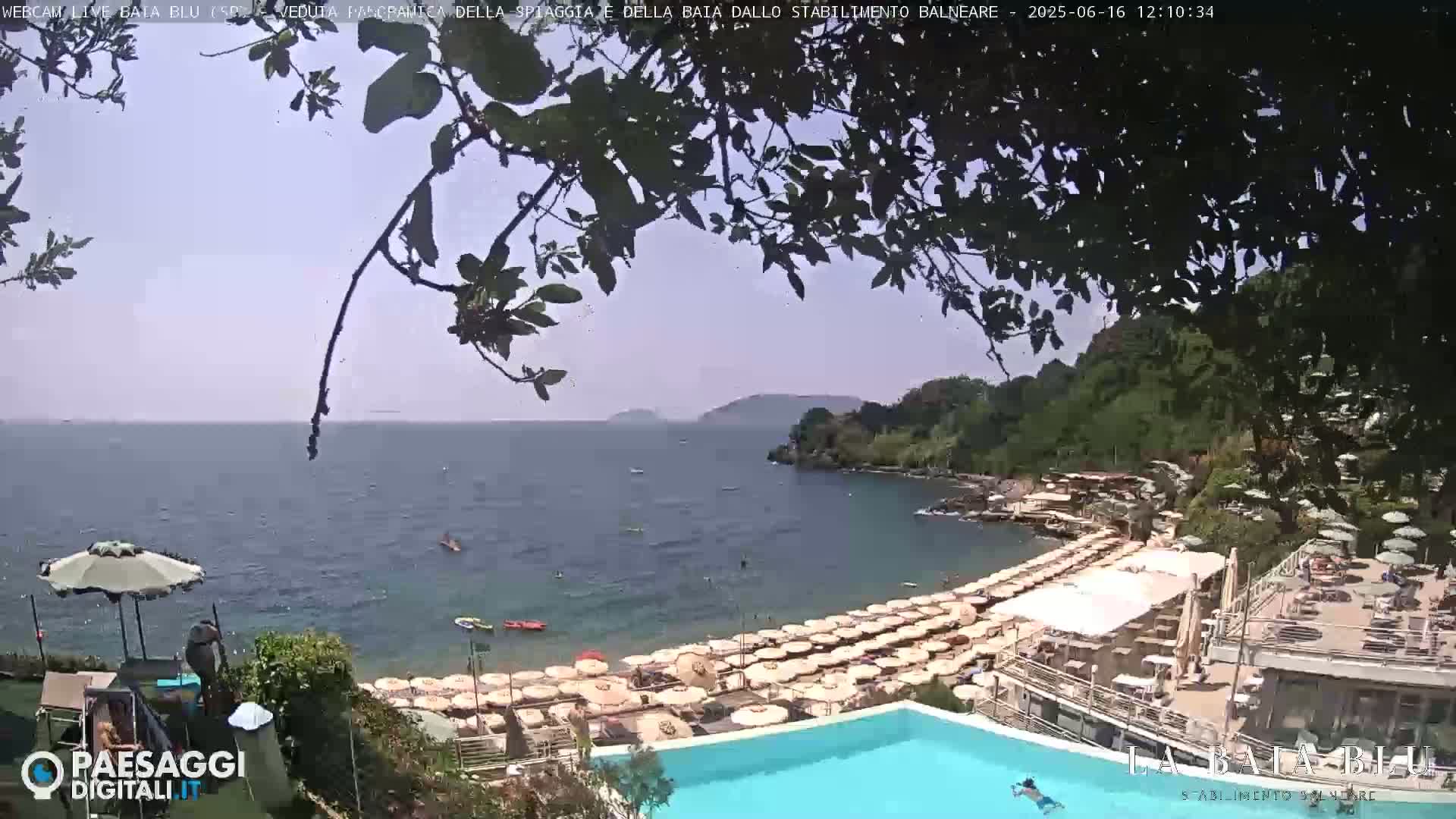 A partly sunny day at a beach club with a swimming pool overlooking a calm bay with numerous beach umbrellas and a few people swimming in the sea.