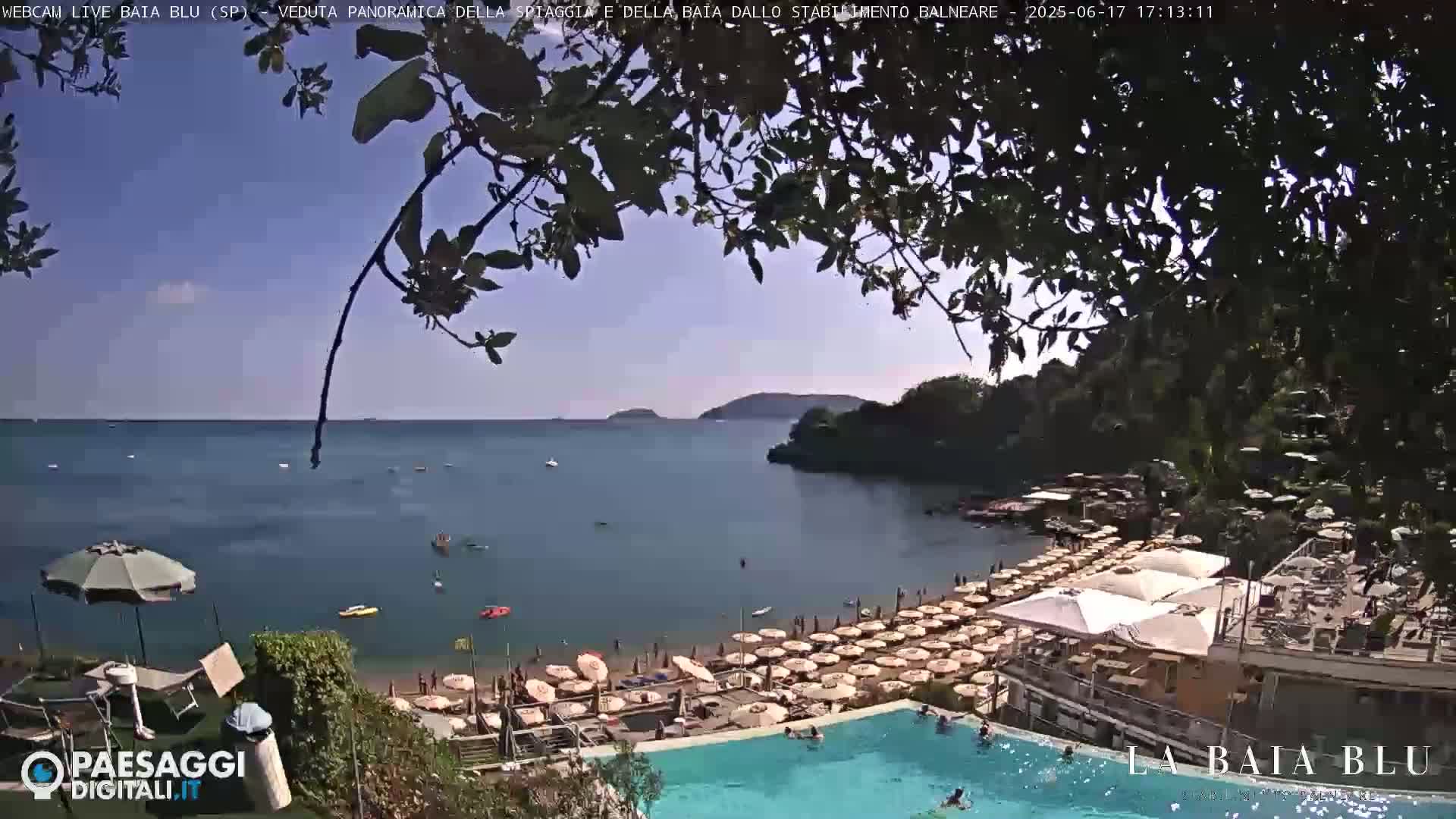A partly sunny view from a hilltop overlooking a calm bay with a crowded sandy beach, numerous beach umbrellas, and a swimming pool.
