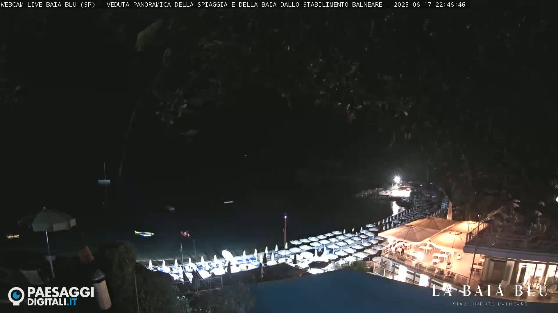 A nighttime, panoramic view of a beach with closed umbrellas and a lit restaurant overlooking a dark bay.