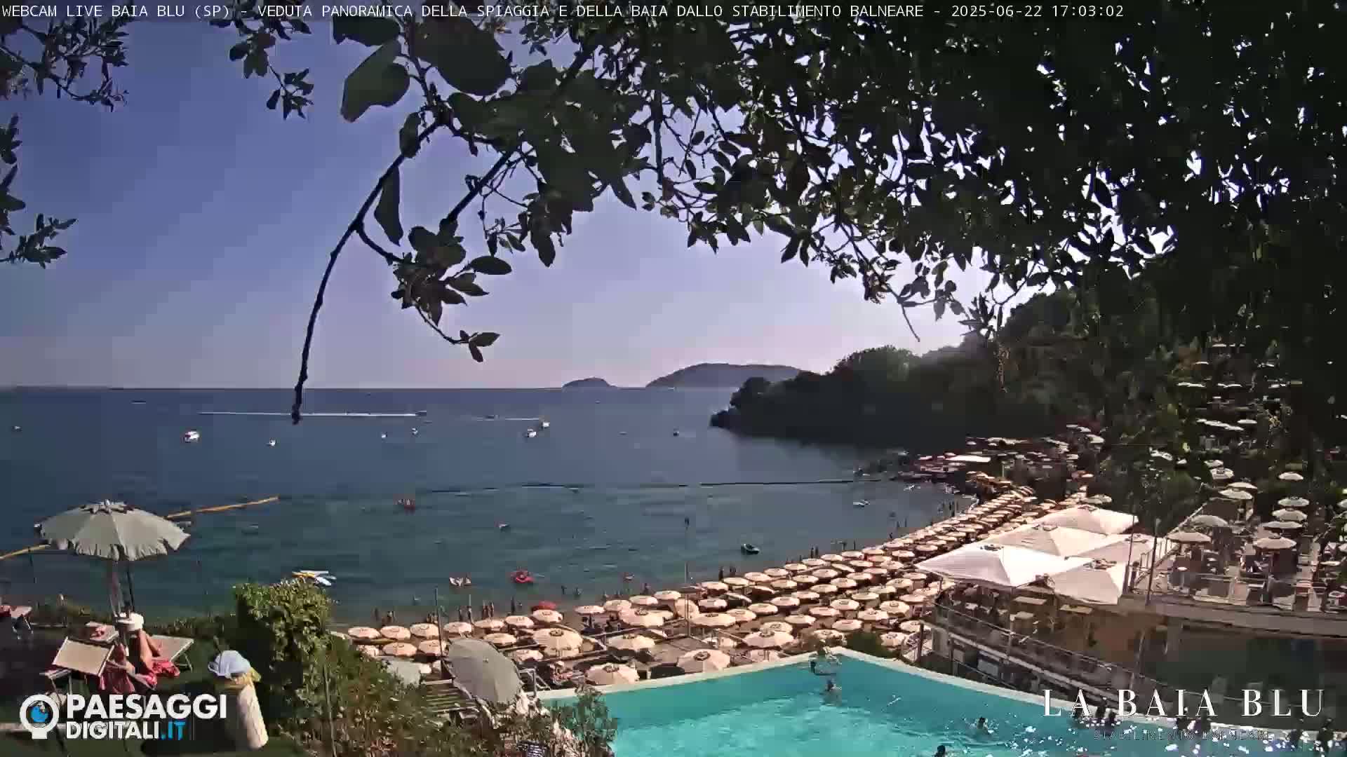 A sunny beach scene shows a calm bay with numerous beach umbrellas and a swimming pool, partially obscured by tree branches.