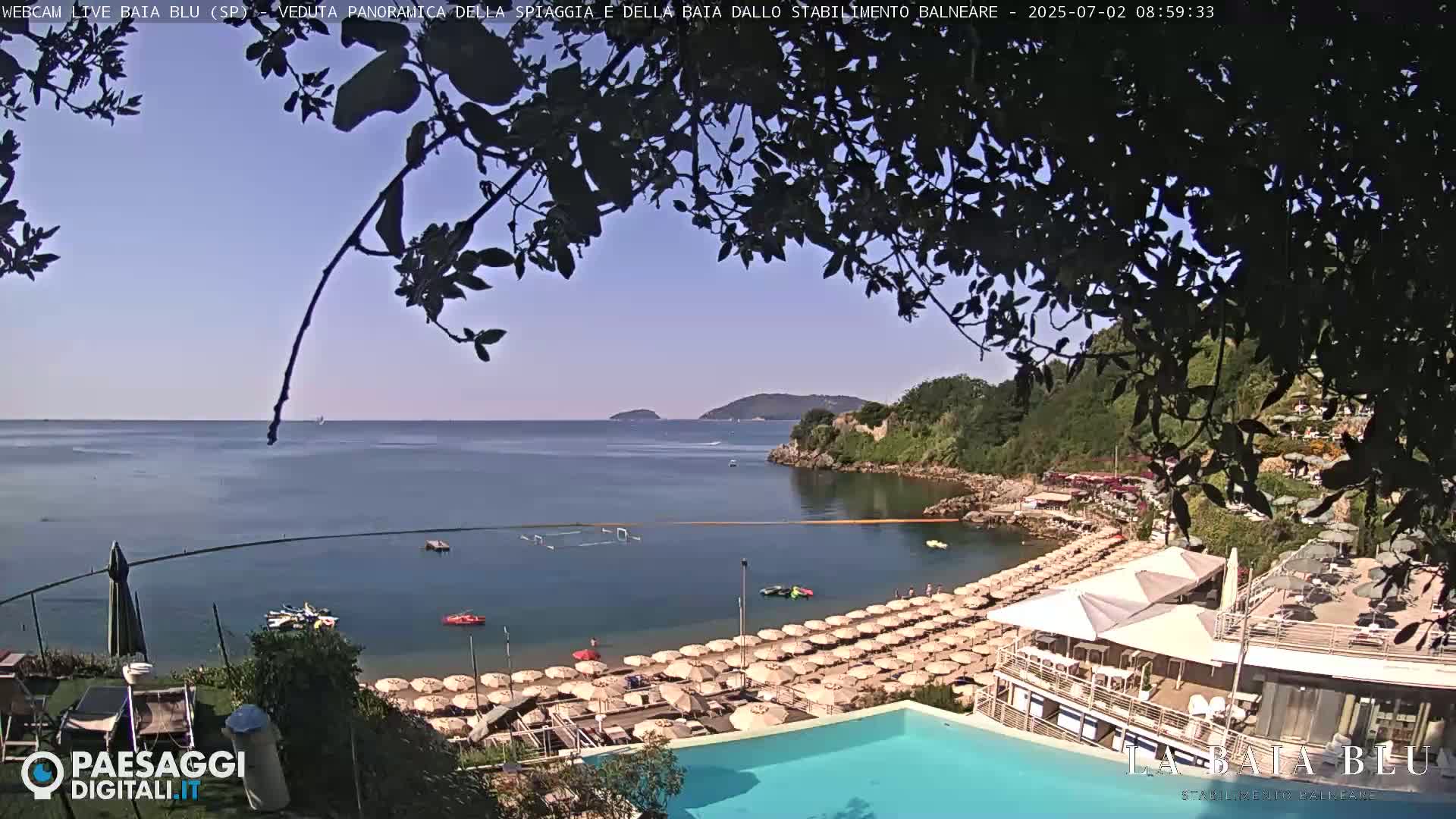A beach with numerous umbrellas and a swimming pool is nestled in a cove, overlooking a calm sea under a clear sky.