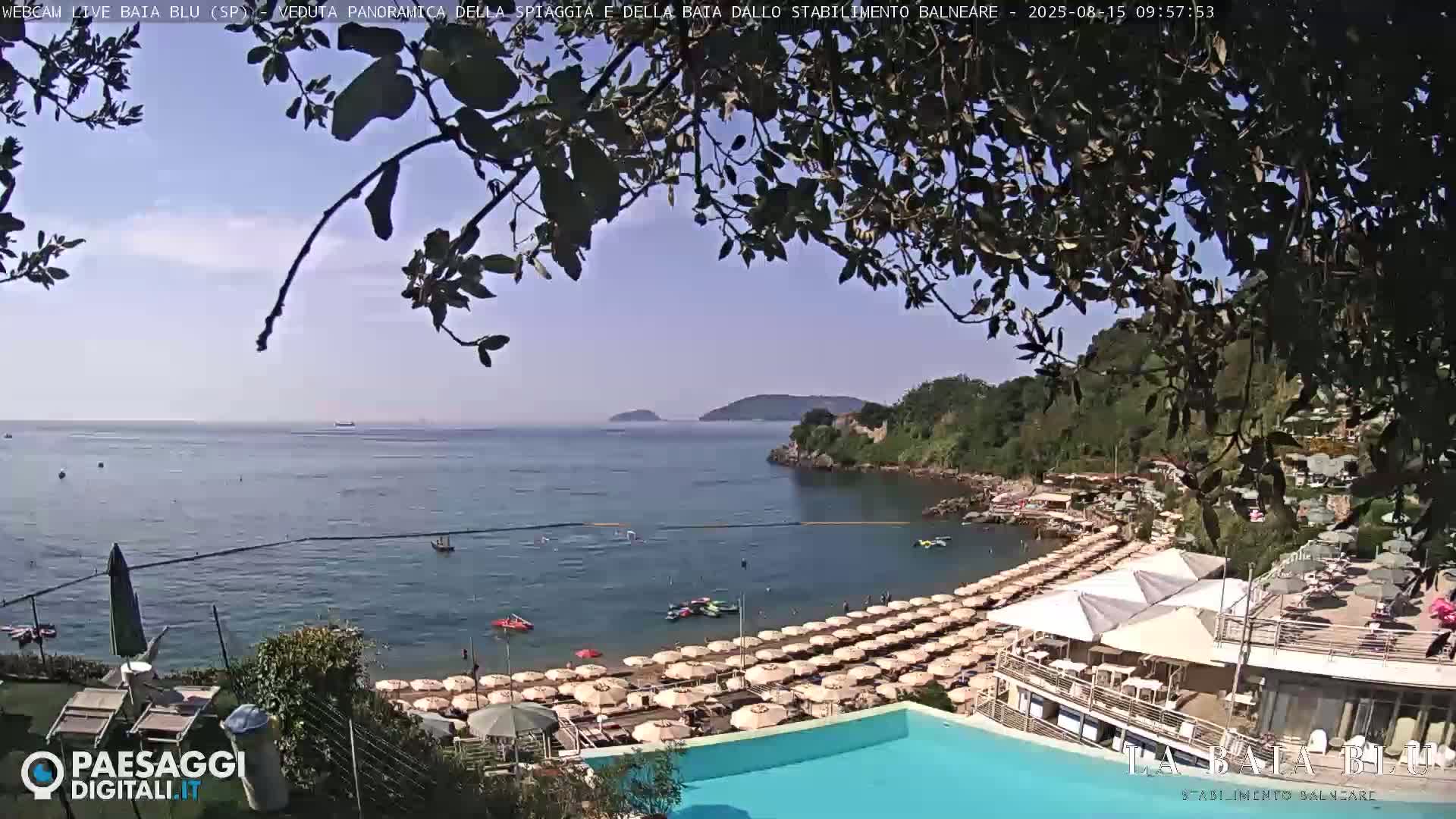 A beach with many umbrellas and a swimming pool is seen from above, overlooking a calm bay and distant hills under a clear sky.