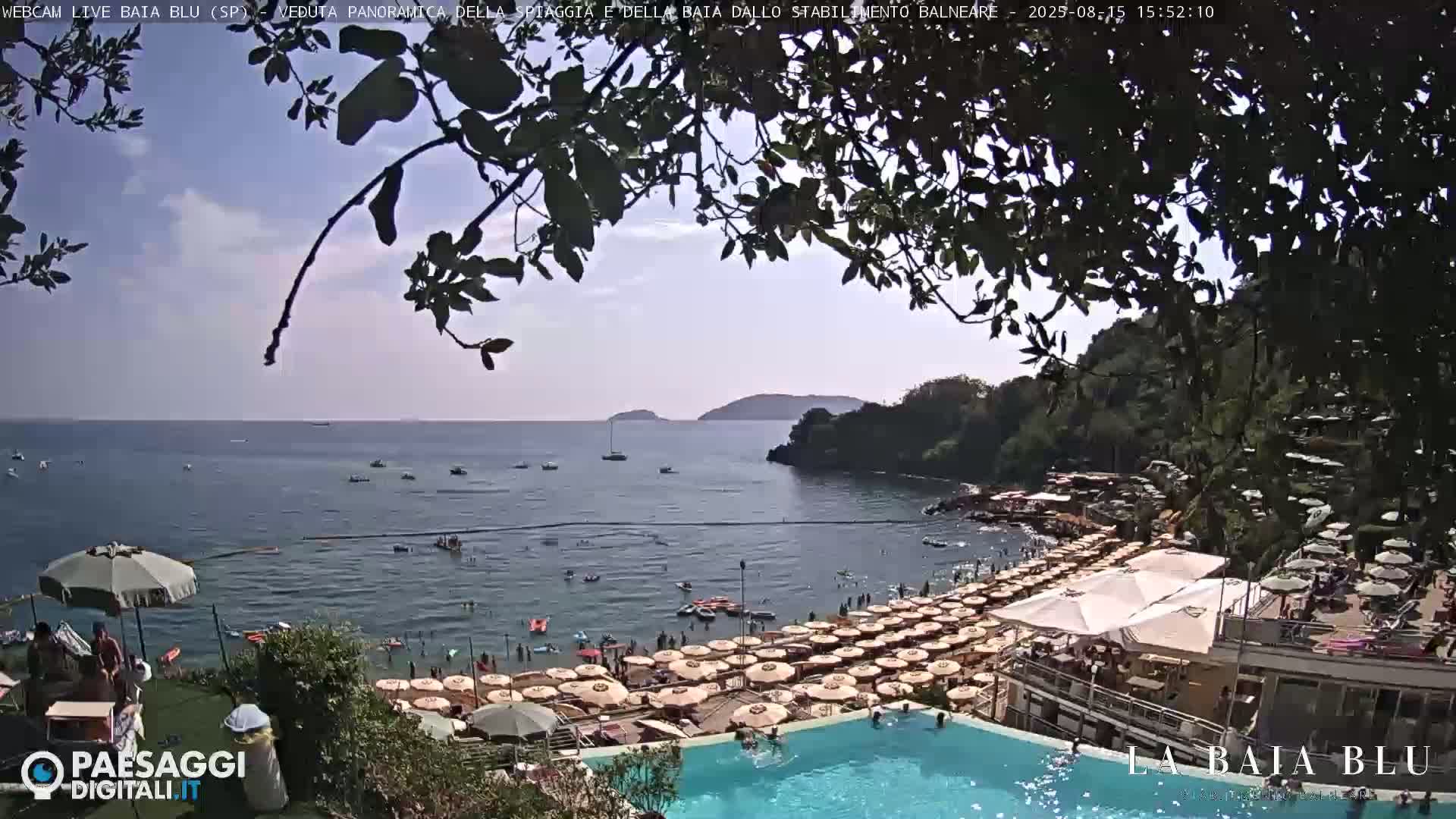 A beach with many umbrellas and a swimming pool is seen from above, overlooking a calm bay and distant hills under a clear sky.