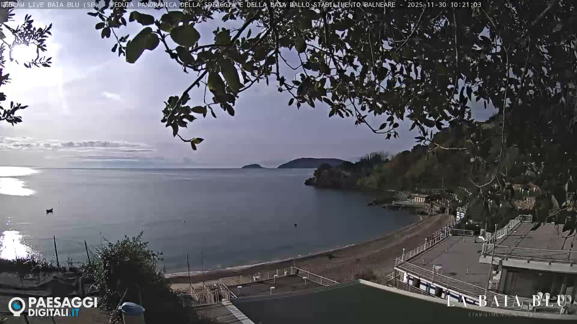 A bright, partly cloudy day reveals a tranquil coastal bay with a pebbly beach, calm blue-gray waters, and distant islands, framed by green foliage and a waterfront building.