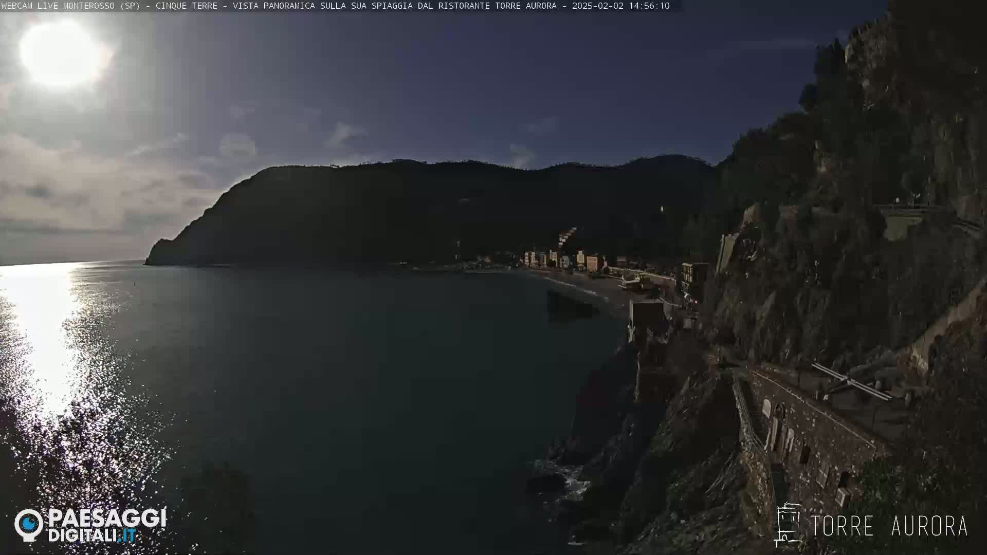 A partly sunny day shows a coastal town nestled between dark hills, with a calm sea reflecting sunlight.