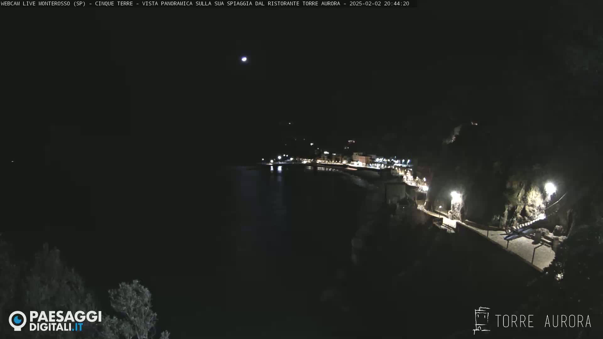 A nighttime, aerial view of a dark, calm body of water next to a dimly lit coastline with buildings and a walkway.