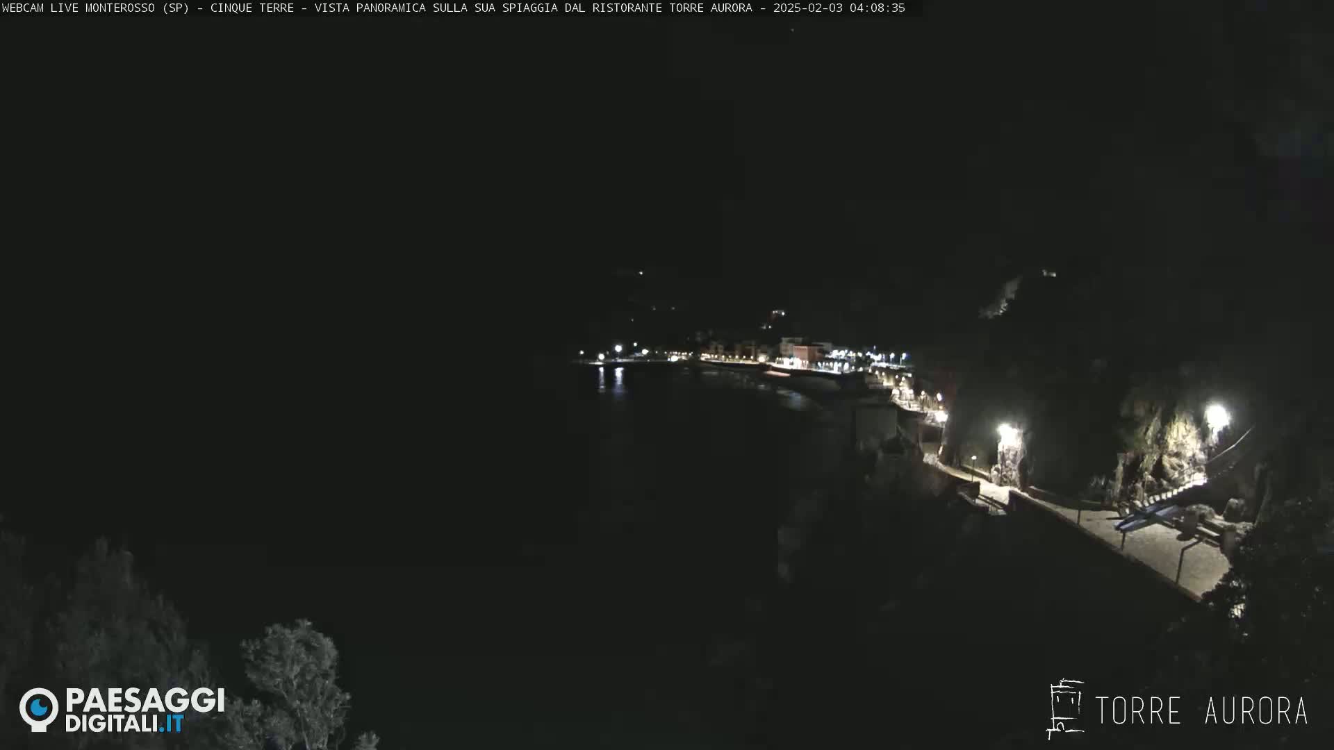 A nighttime, panoramic view of a dark coastline with a few lights visible along a walkway and buildings.