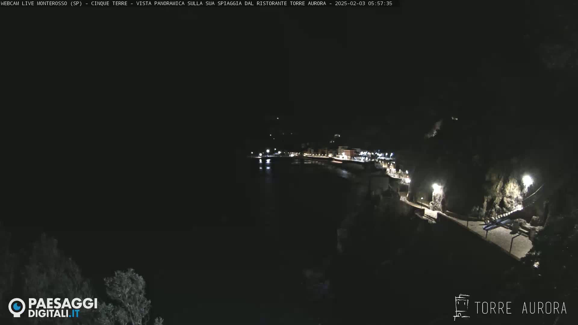 A nighttime, panoramic view of a dark, coastal town with dimly lit buildings and a path along the water.