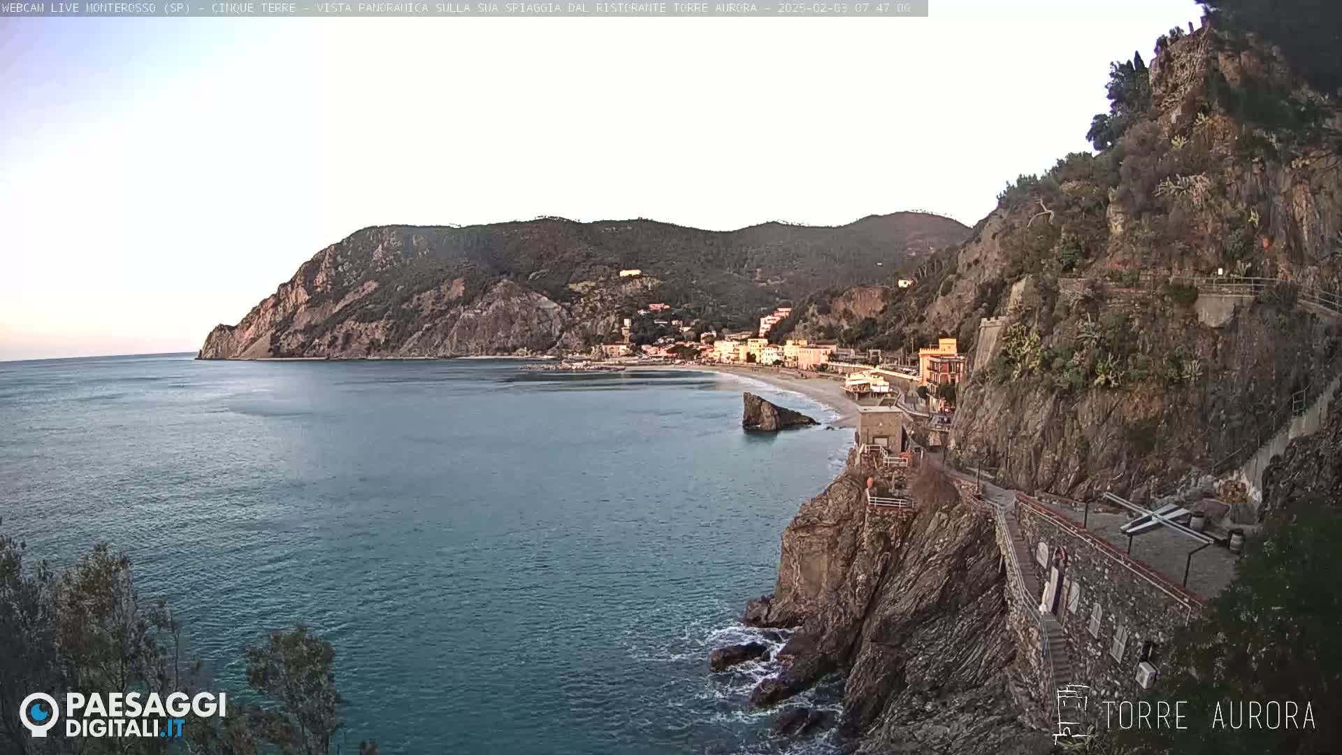 A coastal town nestled between a rocky cliff and the sea, under a clear, light-colored sky.