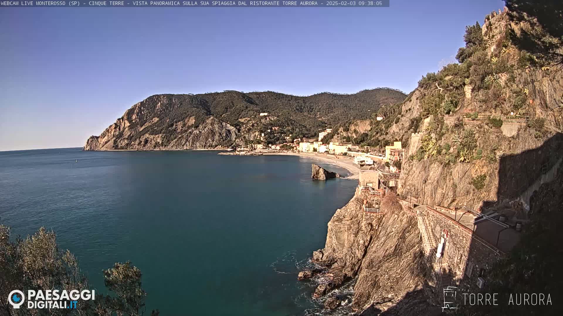 A coastal town nestled between a rocky cliff and the calm, blue sea under a clear, sunny sky.