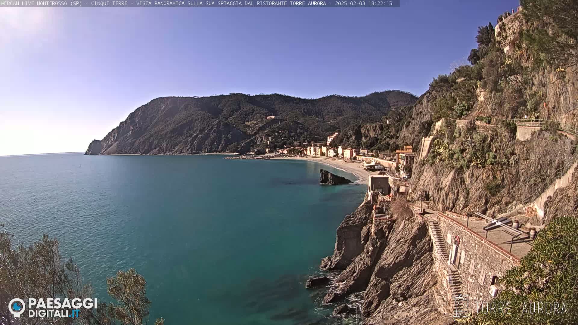 A sunny coastal scene shows a town nestled between teal water and steep, rocky cliffs with a staircase leading up the cliffside.