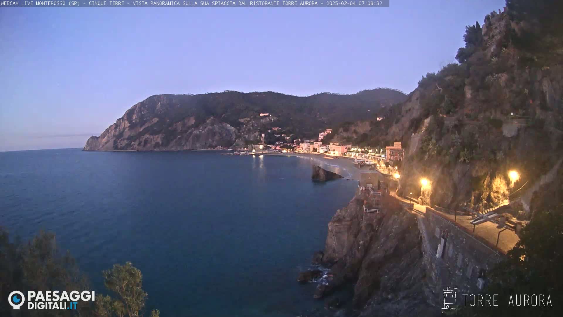 A coastal town nestled between dark hills and a calm, dark blue sea under a twilight sky.