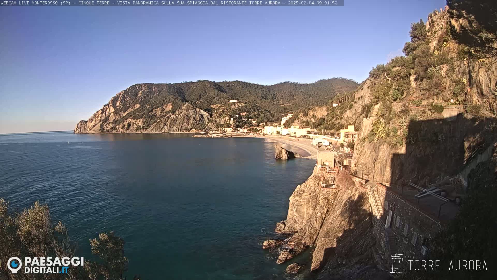 A coastal town nestled between steep, rocky cliffs overlooks a calm, blue sea under a clear, sunny sky.