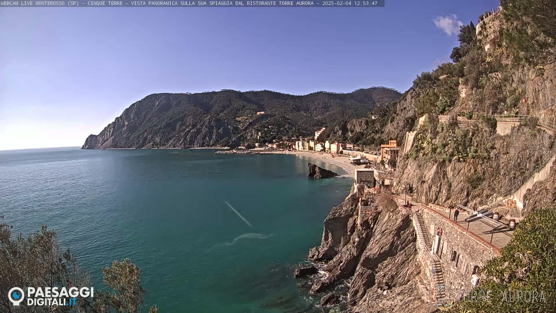 A coastal town nestled between rocky cliffs and a calm, teal sea under a clear blue sky.