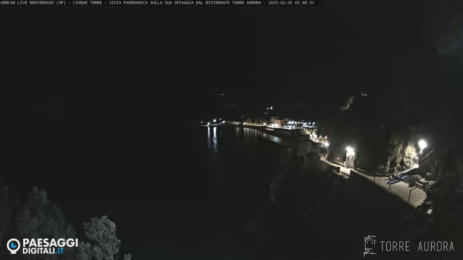 A nighttime, aerial view shows a dark coastline with illuminated buildings and a walkway along the water.