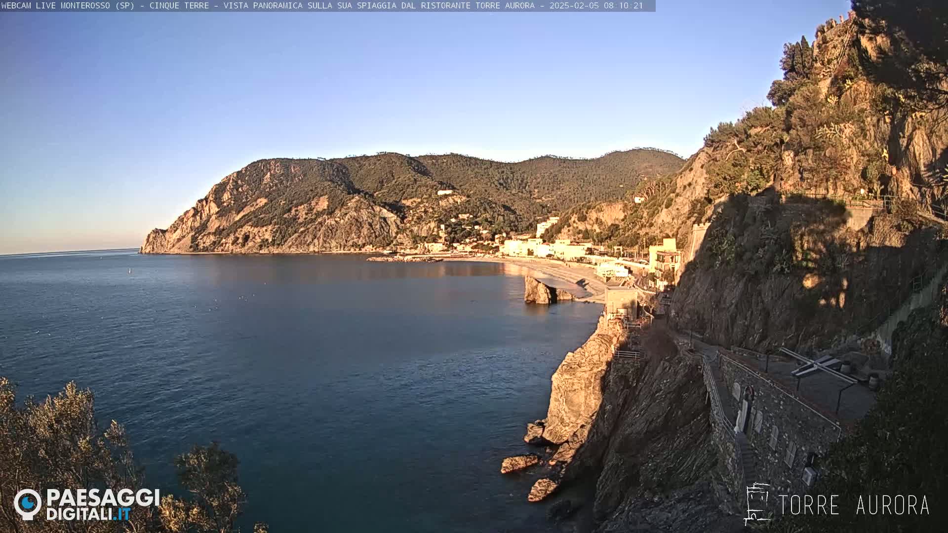 A coastal town nestled between a rocky cliff and the calm, blue sea under a clear, bright sky.