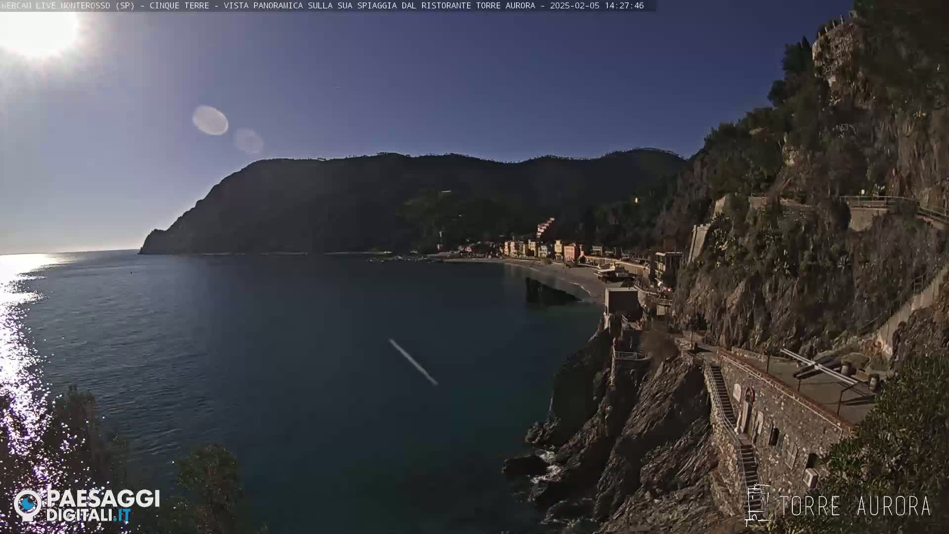 A sunny day reveals a coastal town nestled between dark green hills and a calm blue sea.