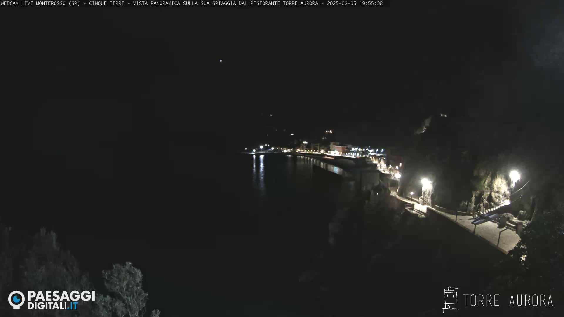A nighttime, panoramic view of a coastal town with a dark, calm body of water, illuminated by lights along a walkway and buildings.