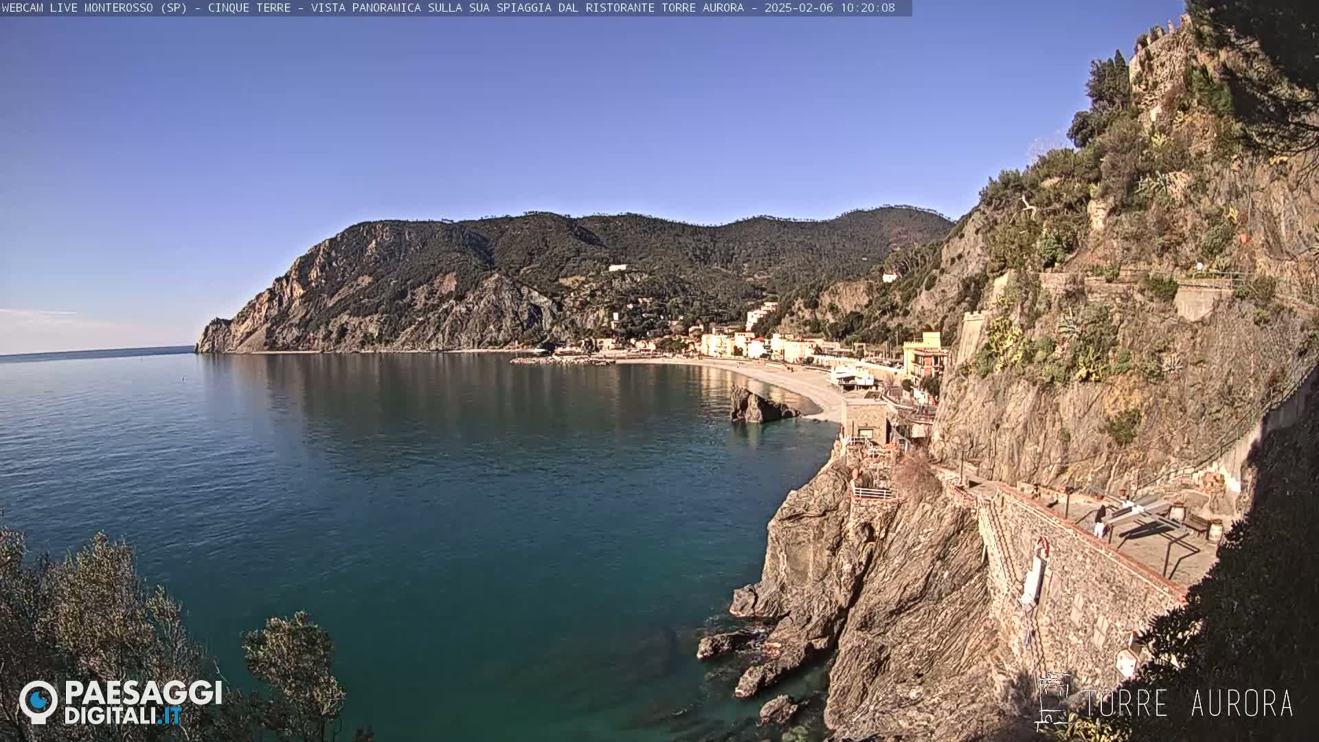 A clear, sunny day reveals a coastal town nestled between a rocky cliff and the calm, dark-blue sea.
