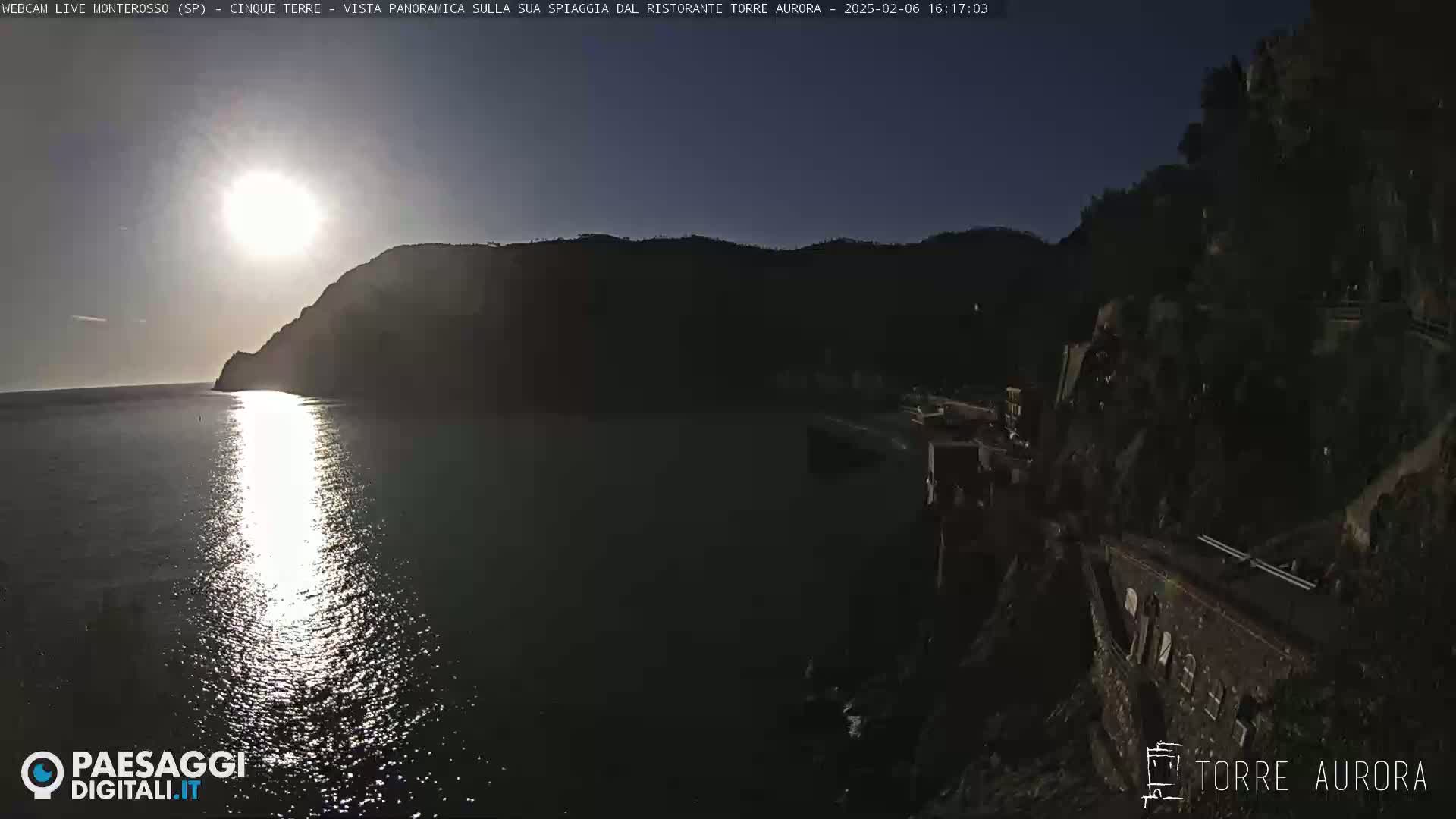 A bright sun shines over a calm body of water reflecting its light, nestled between dark hillsides on a clear day.