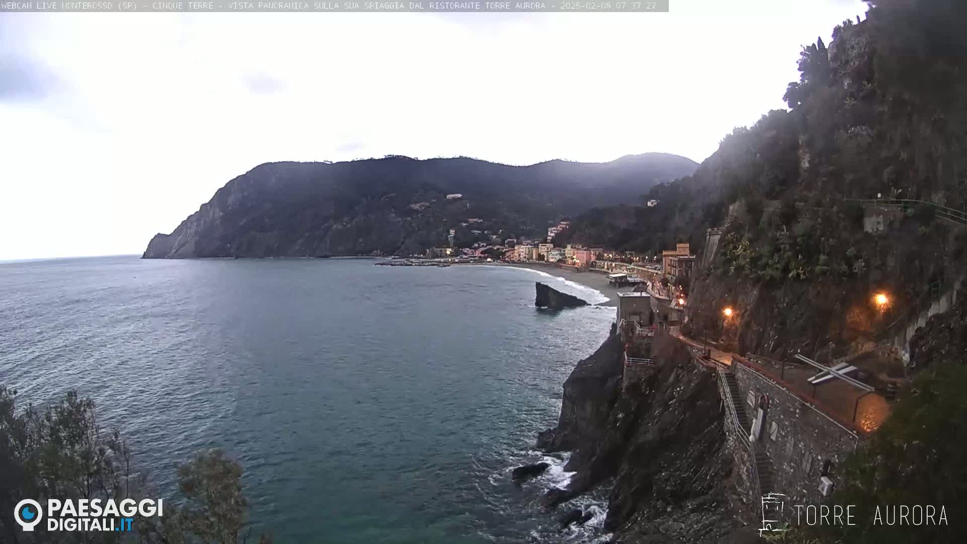 A coastal town nestled between dark, rocky cliffs overlooks a calm sea under an overcast sky.