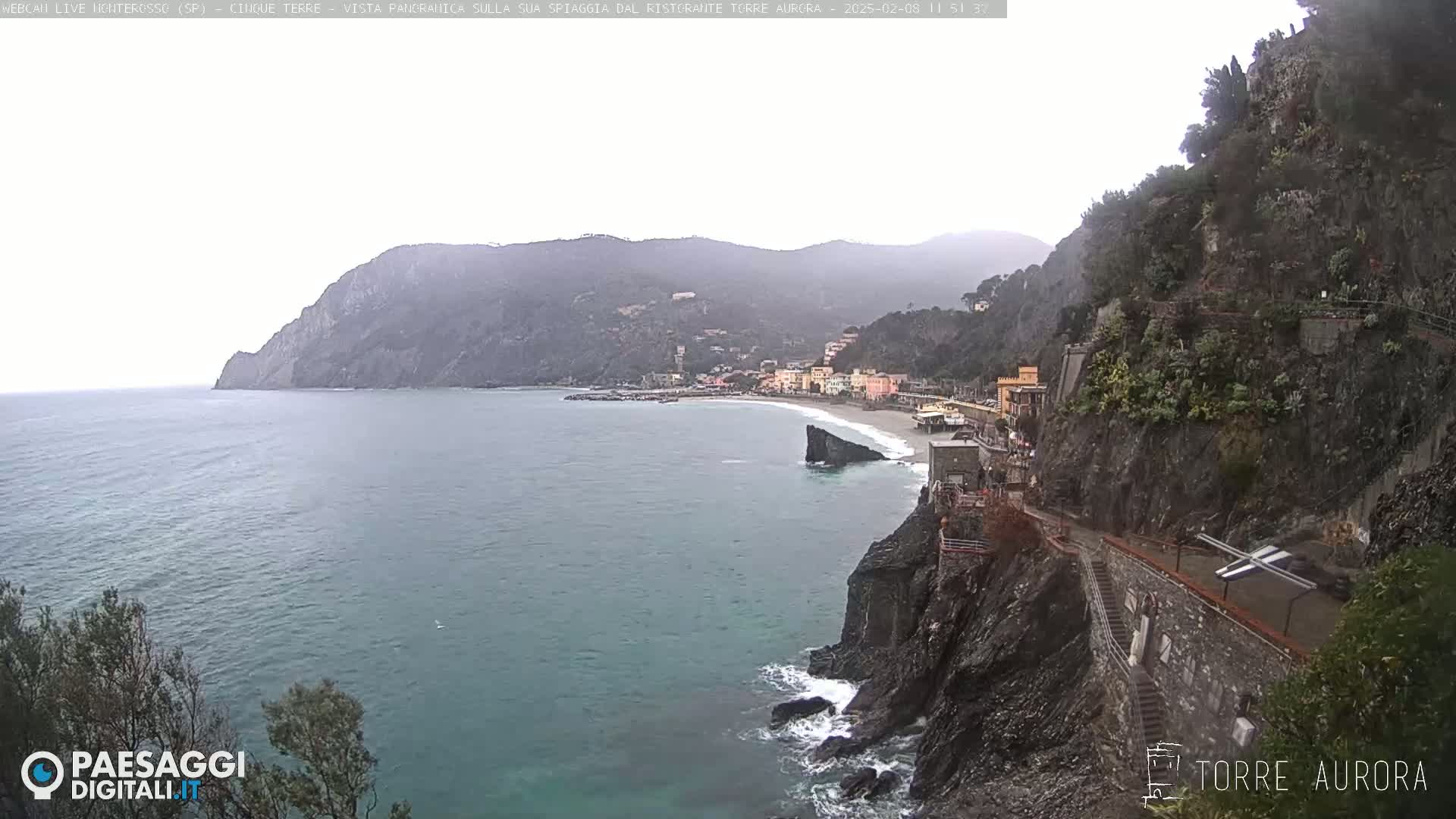 A coastal town with a sandy beach nestled between steep, rocky cliffs is seen on an overcast day.