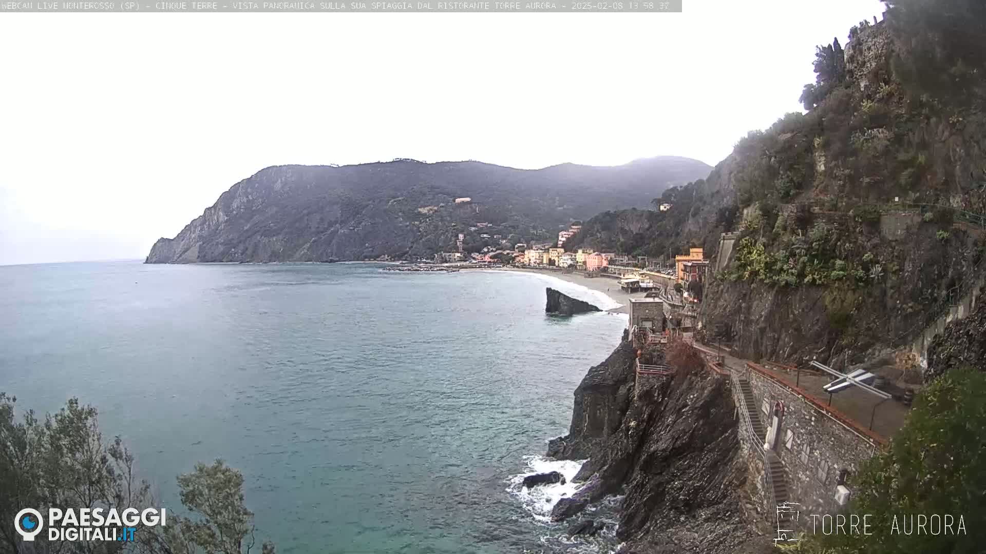 A coastal town nestled between steep, rocky cliffs overlooks a calm sea under an overcast sky.