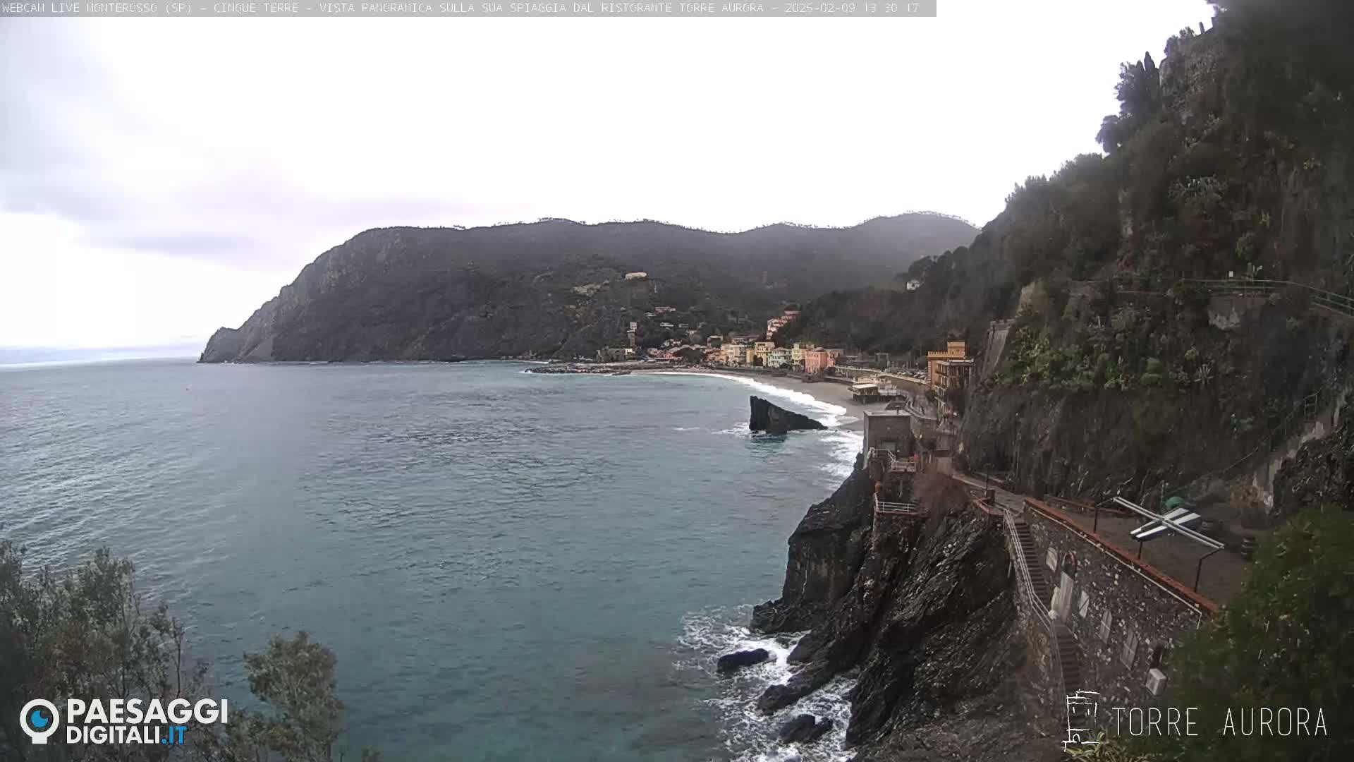 A coastal town nestled between steep, dark cliffs and a calm, blue-green sea under an overcast sky.