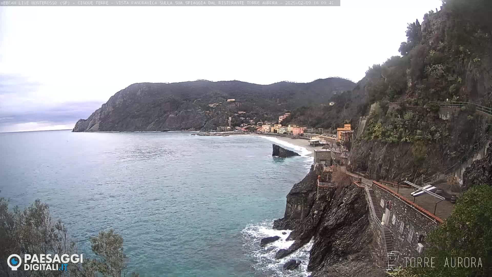 A coastal town nestled between hills overlooking a calm sea under an overcast sky.