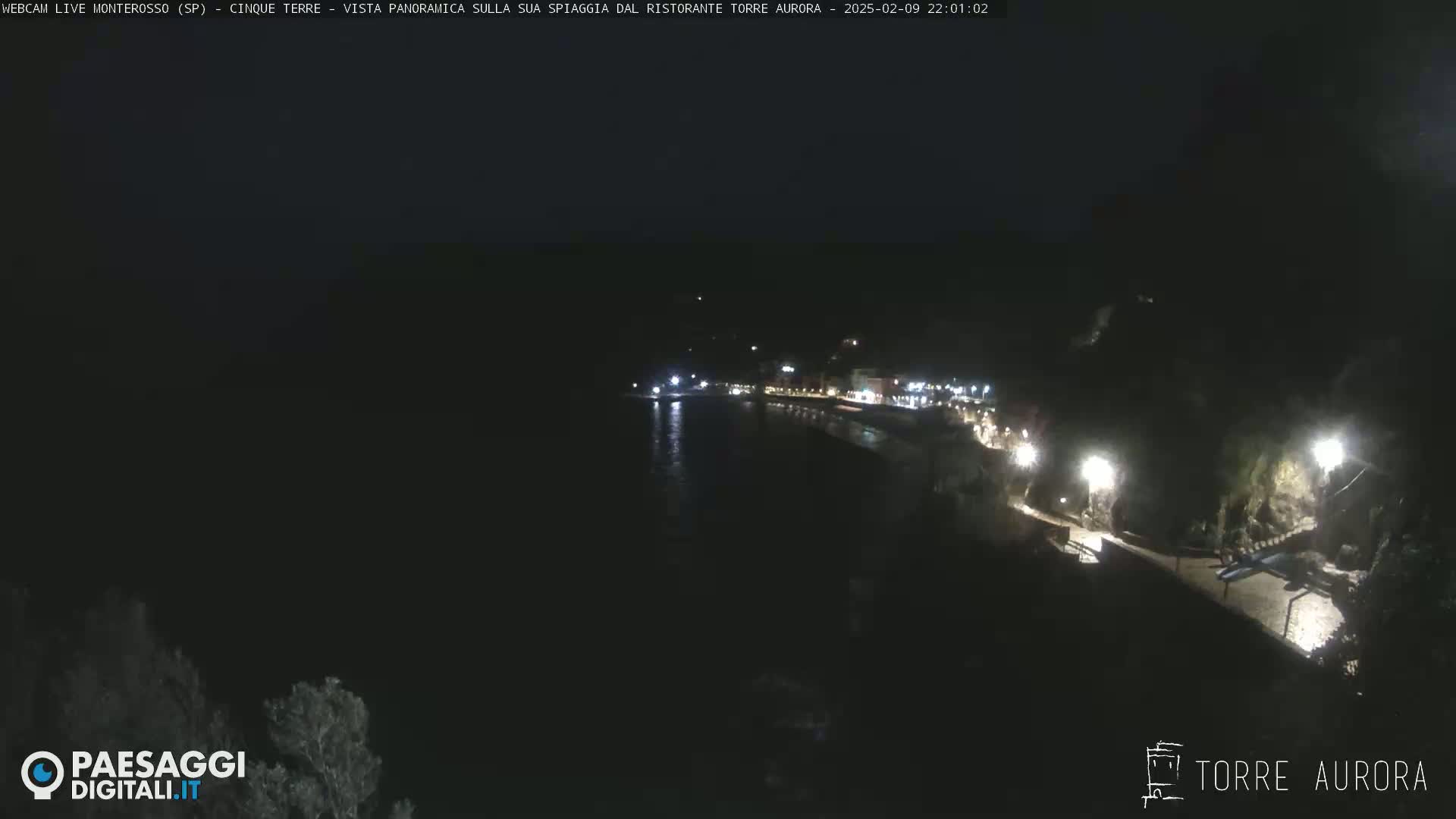 A nighttime, panoramic view of a dark coastline with dimly lit buildings and a walkway along the water's edge.