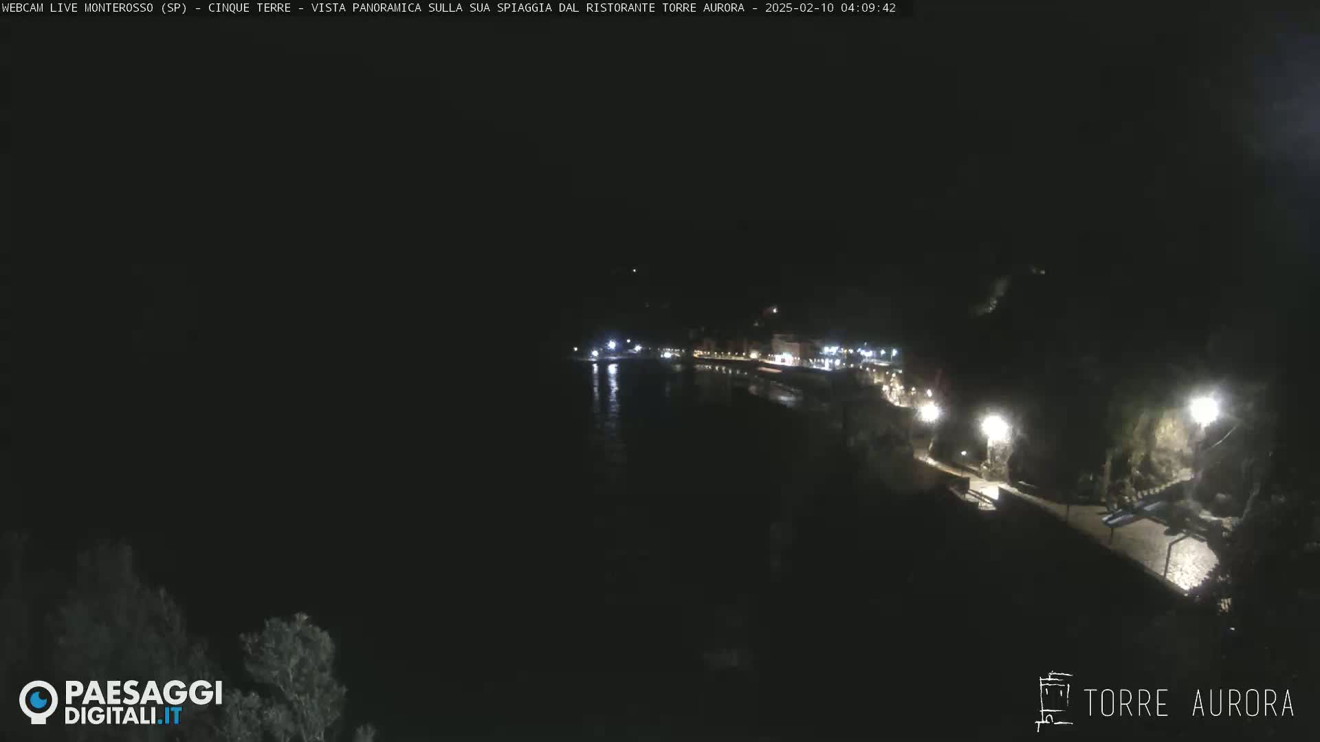 A nighttime, long shot of a dark coastline with a few lights illuminating buildings and a walkway along the water.