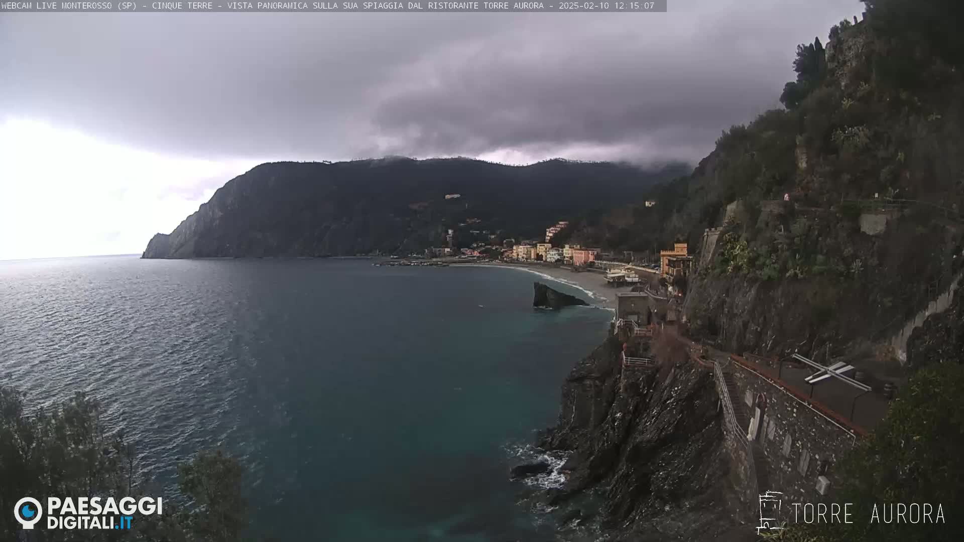 A coastal town nestled between dark, rocky cliffs and a calm, dark-blue sea under an overcast sky.