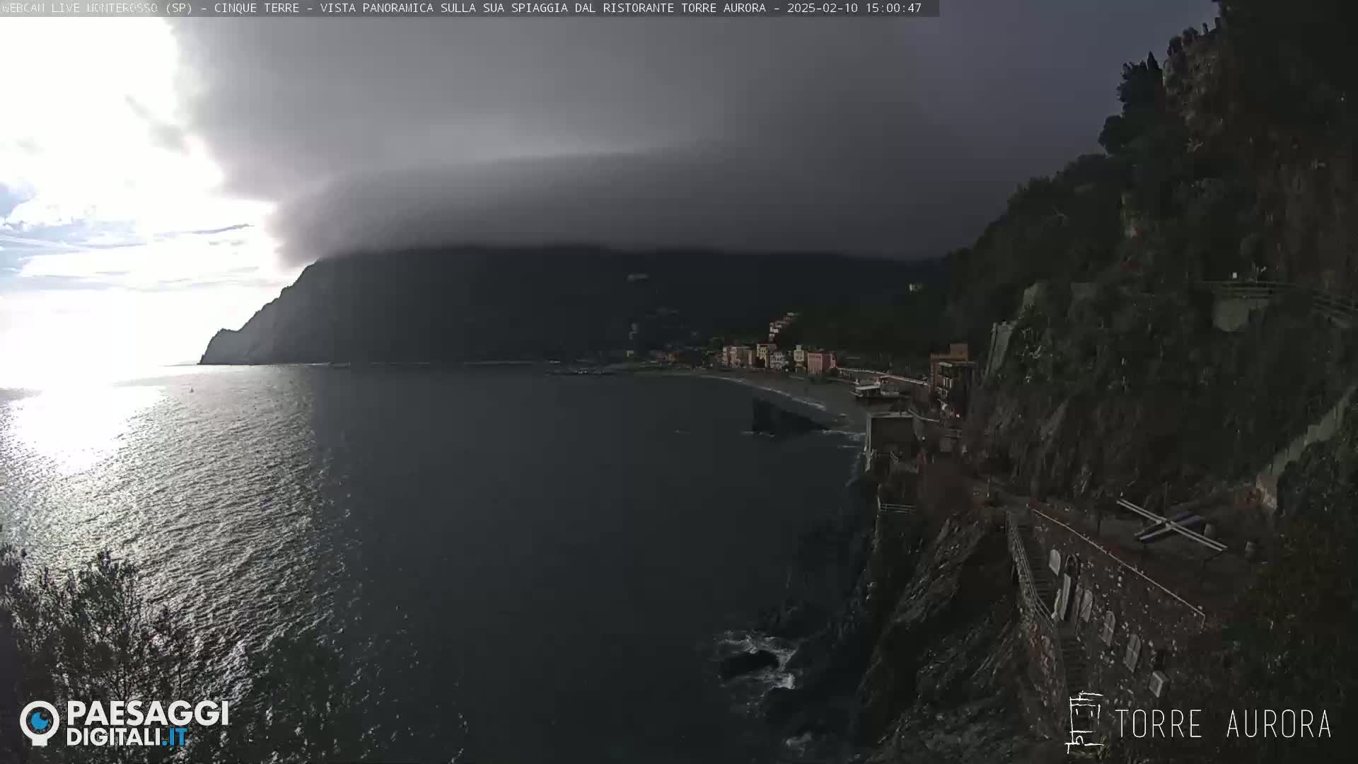 A dark, overcast sky hangs over a coastal town nestled between dark cliffs and a calm sea.