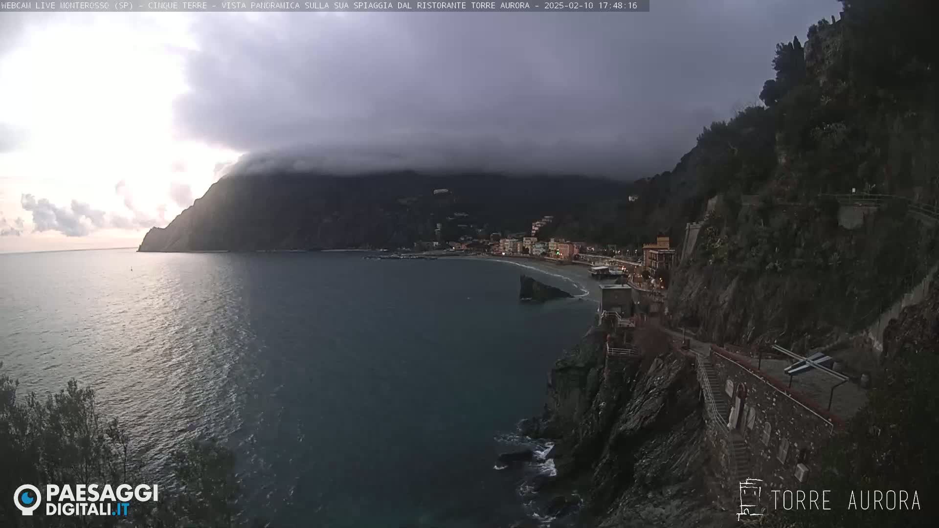 A coastal town nestled between dark, rocky cliffs is partially obscured by low-hanging clouds under an overcast sky.