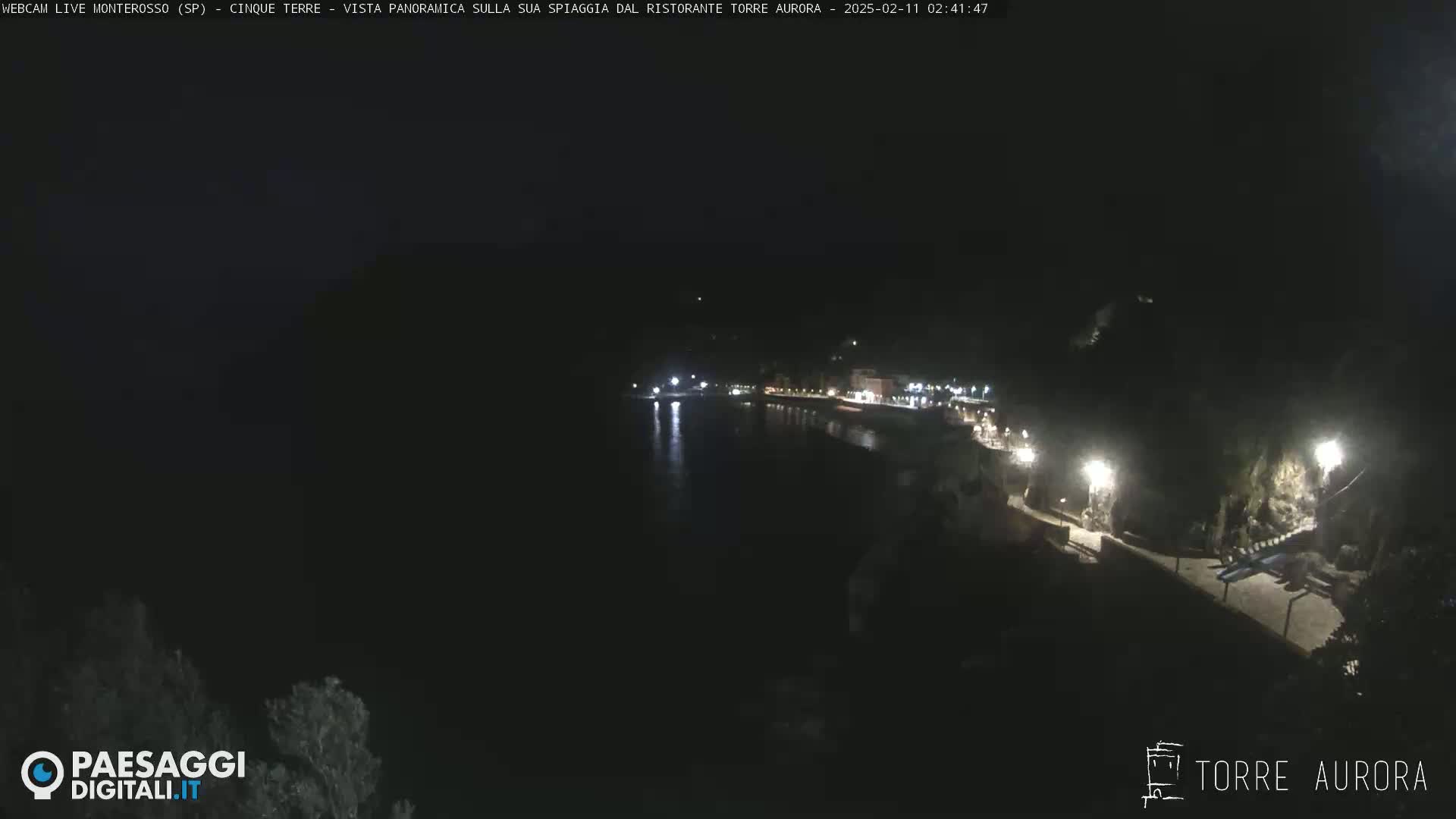 A nighttime, aerial view of a dimly lit beach and coastline, with calm water reflecting the lights.