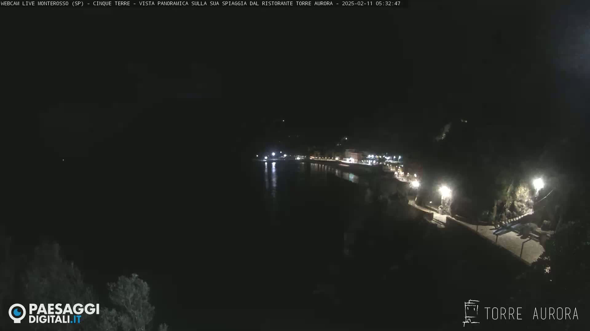 A nighttime, aerial view shows a dark, calm body of water next to a dimly lit coastline with buildings and paths.