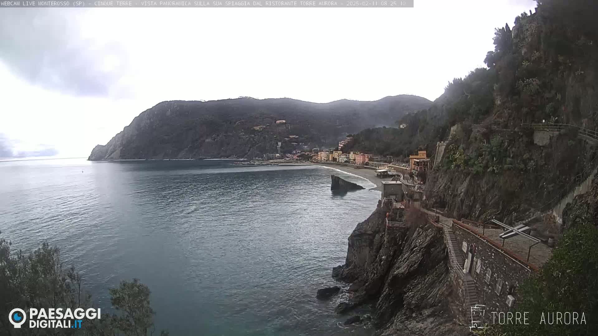 A coastal town nestled between a steep, rocky cliff and a calm, gray sea under an overcast sky.