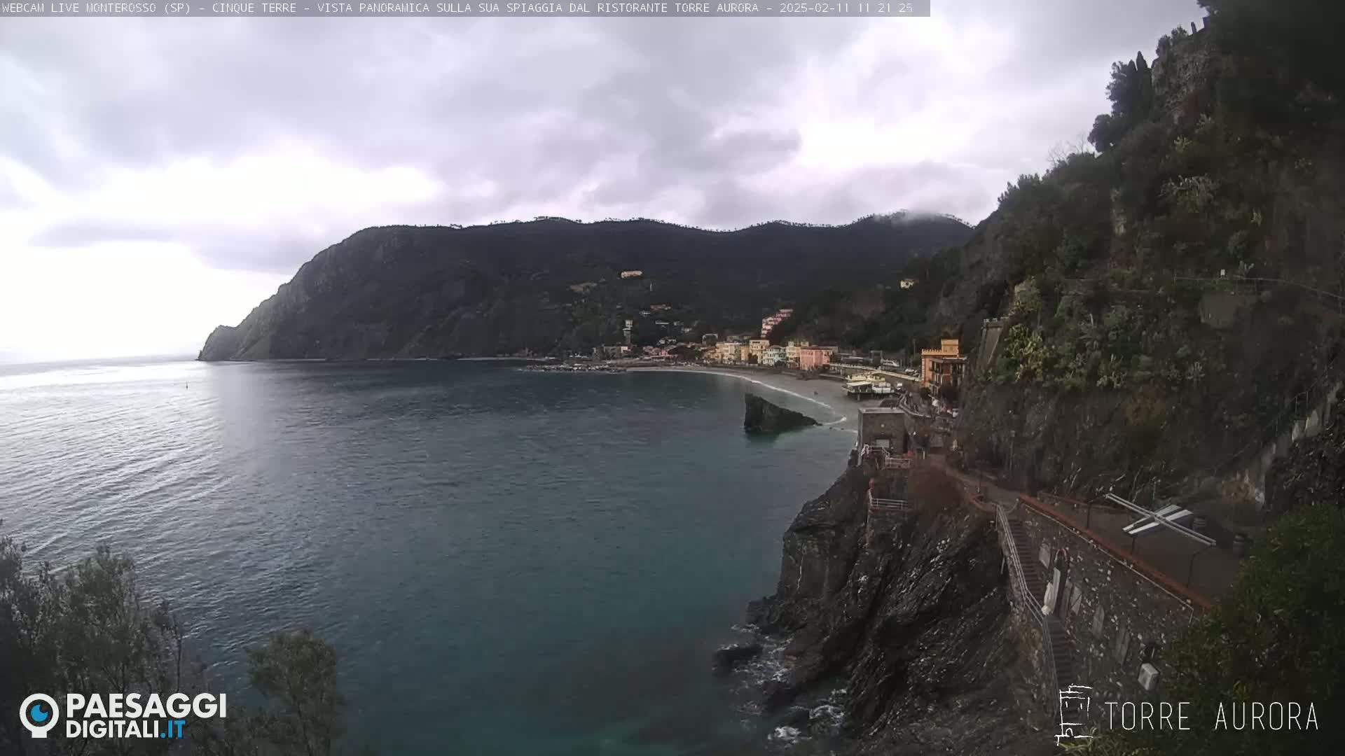 A coastal town nestled between dark, rocky cliffs on a cloudy day, with a calm, dark-blue sea stretching to the horizon.
