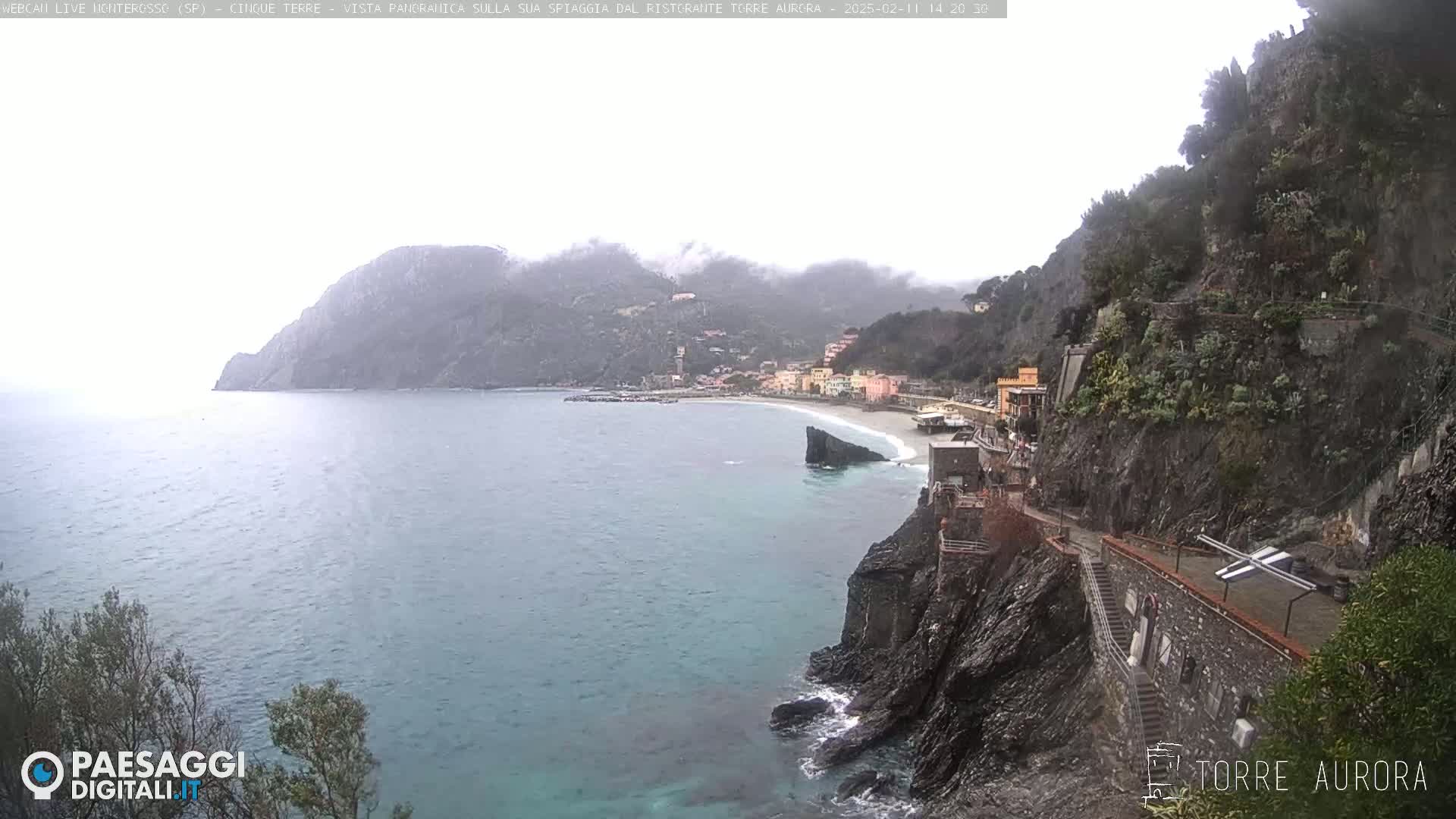 A coastal town with a beach is partially obscured by fog and mist, seen from a high vantage point overlooking a rocky cliffside.