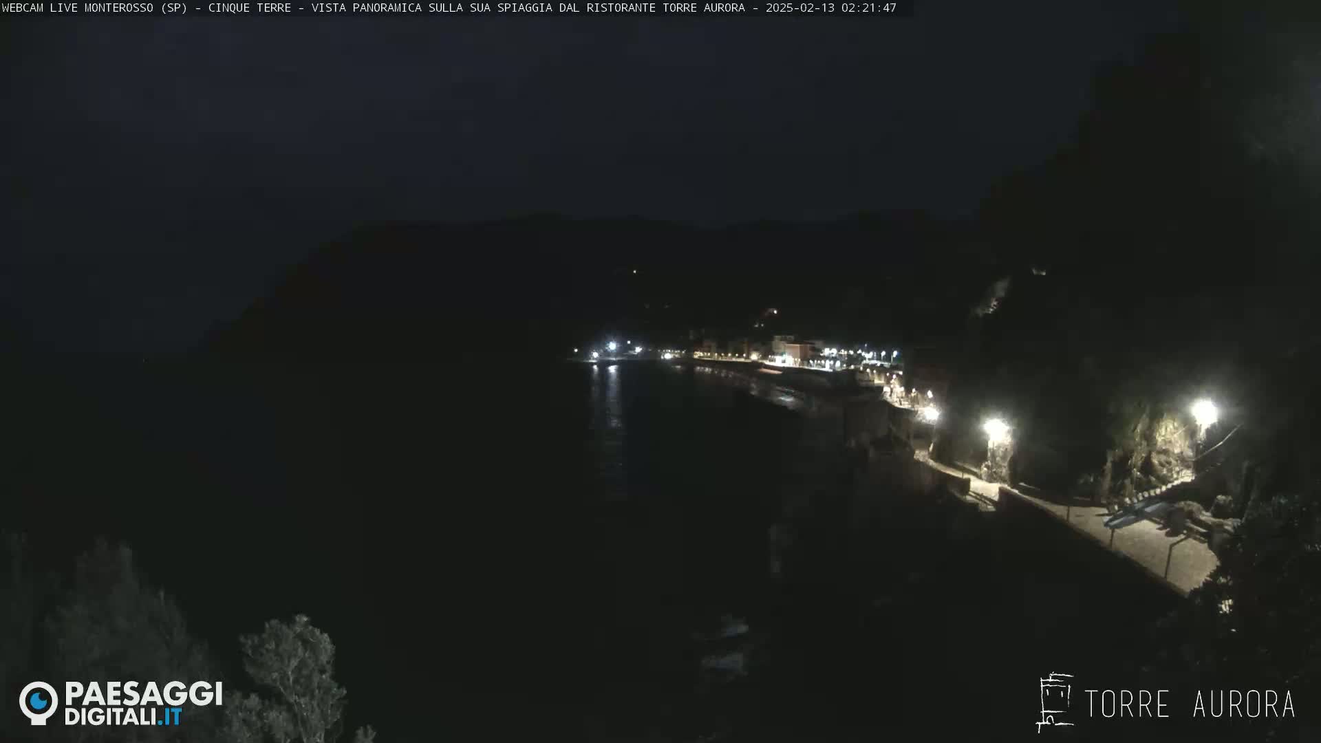 A nighttime, panoramic view of a dark coastline with a few lights illuminating a walkway and buildings along the water.