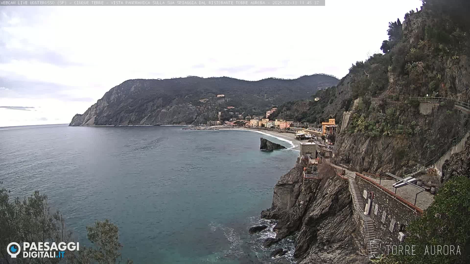 A coastal town nestled between steep cliffs and a calm, blue-green sea under a mostly cloudy sky.