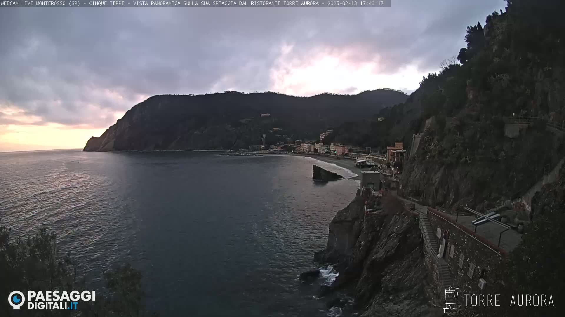 A coastal town nestled against a steep, dark hillside overlooks a calm sea under a cloudy, dusky sky.